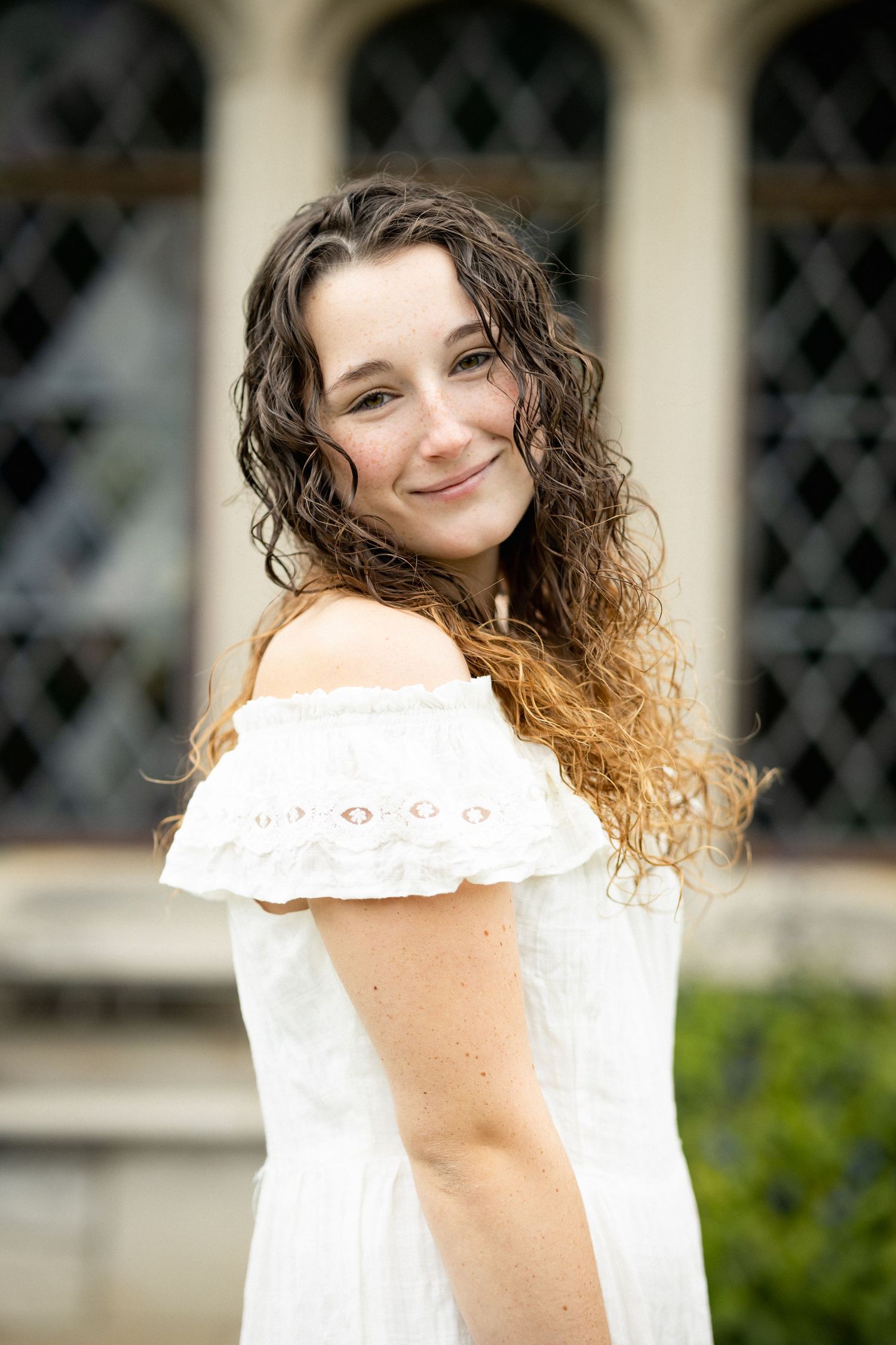 Smiling young woman with curly hair wearing white off-shoulder dress posing before an elegant building.
