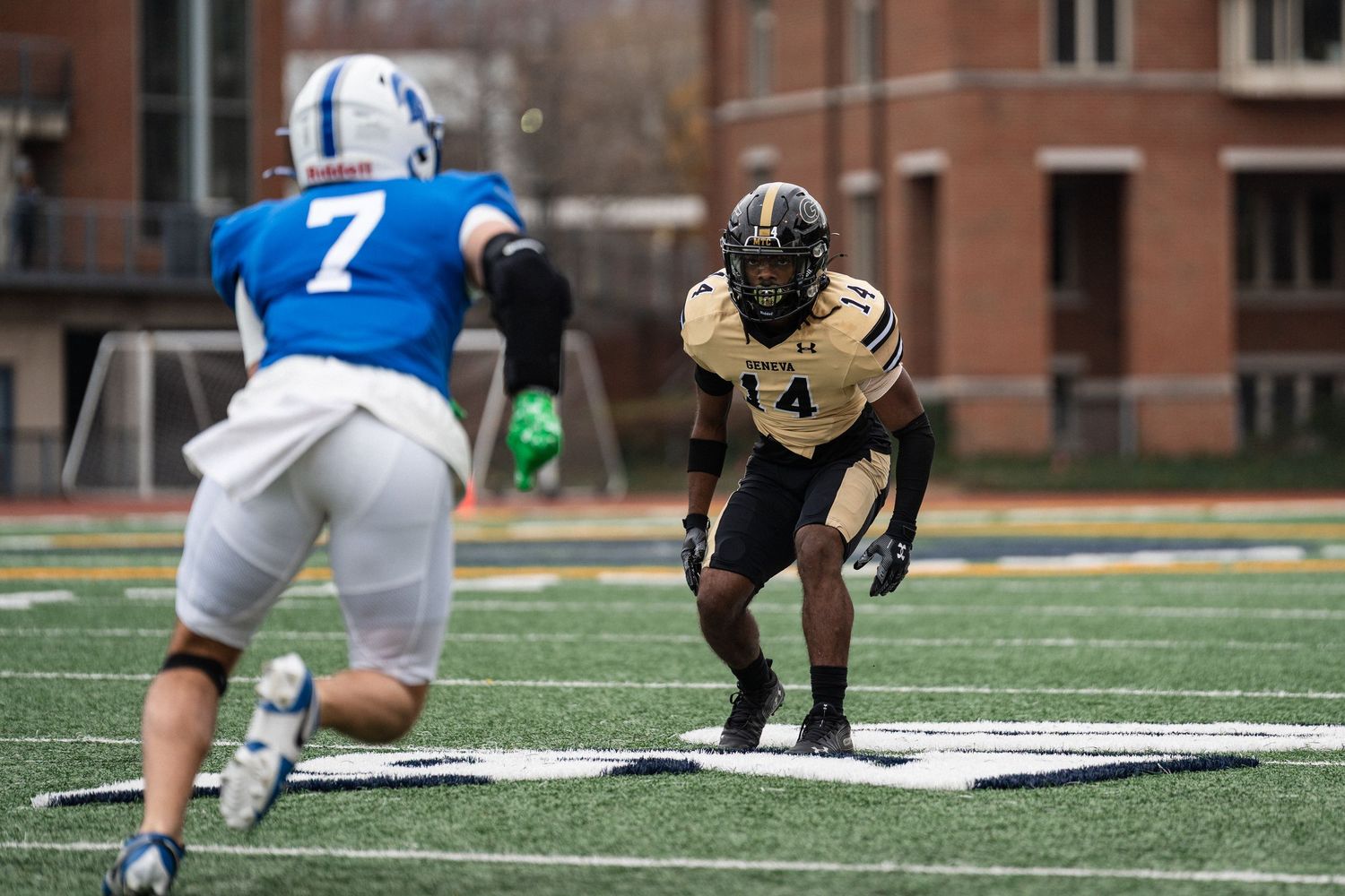 Geneva College defender number 14 in gold uniform squares up against blue-uniformed receiver.