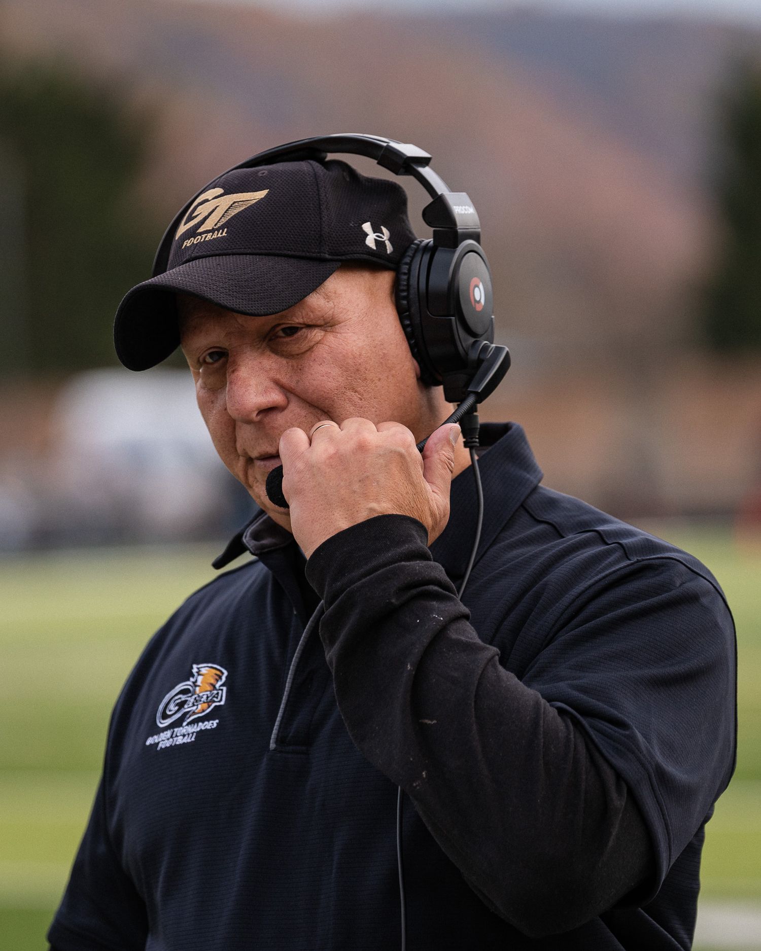 Football coach wearing headset and black jacket on sideline during game.