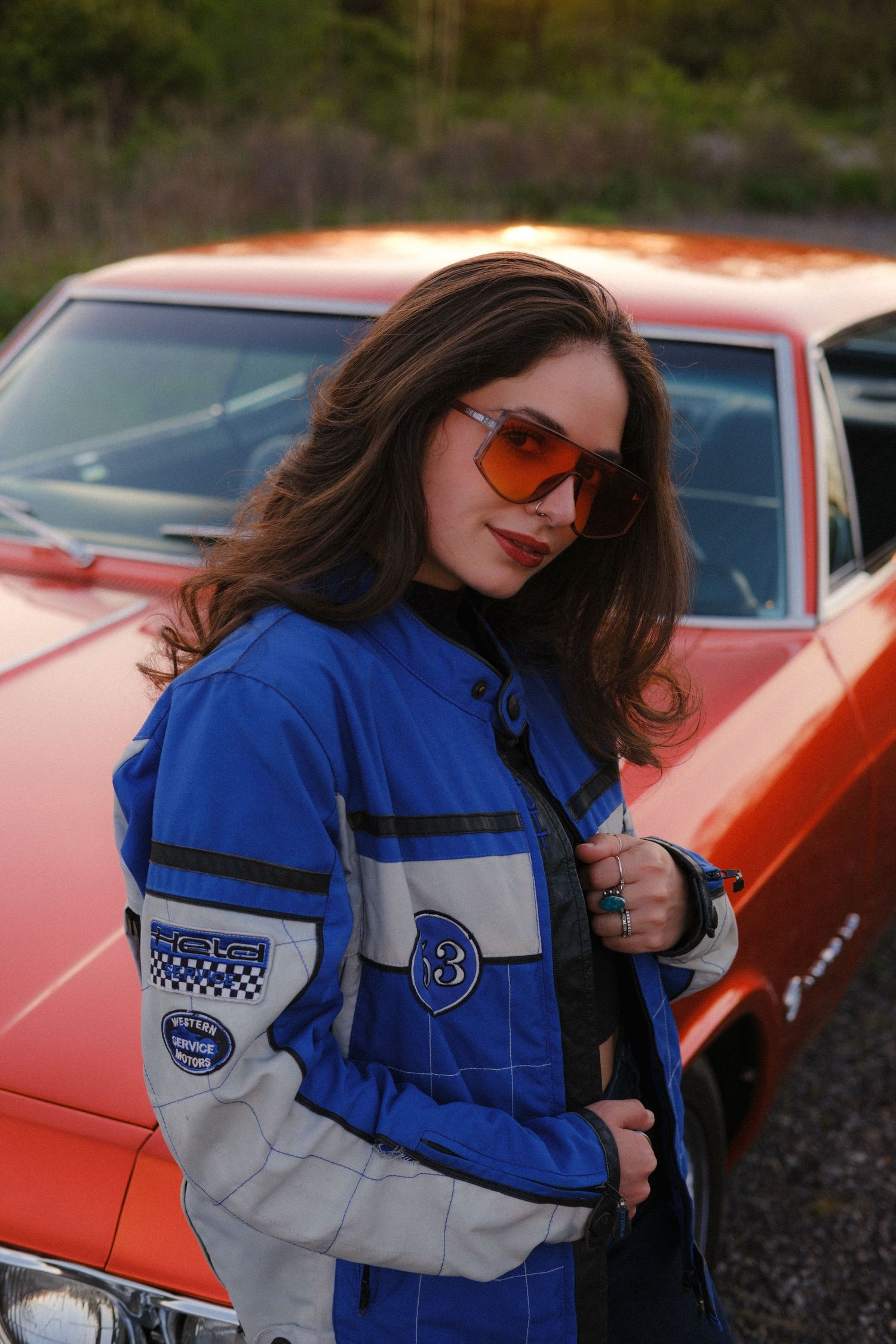 Young woman in blue racing jacket and orange tinted sunglasses standing beside a vintage red muscle car.
