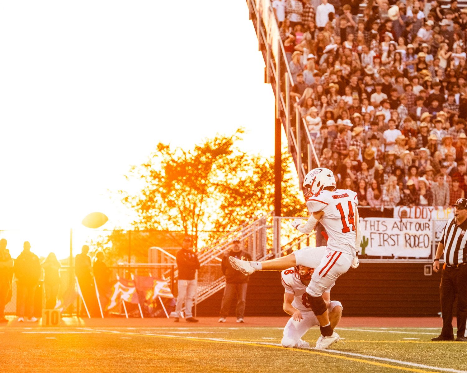 Football player kneeling on sideline at sunset during a high school game with packed stadium crowd in background.