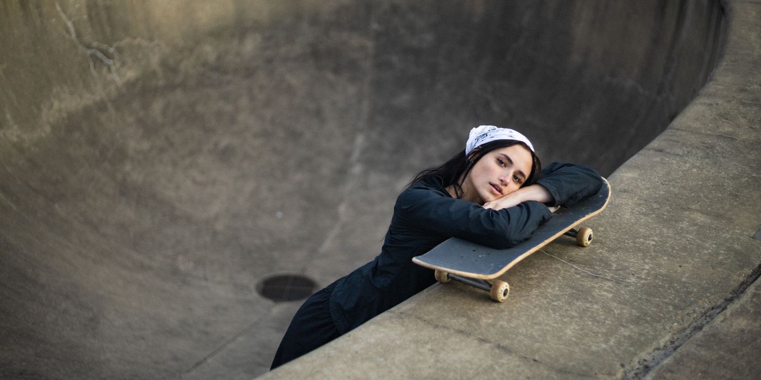 Young woman resting on skateboard in concrete skate park bowl wearing dark hoodie and white beanie.