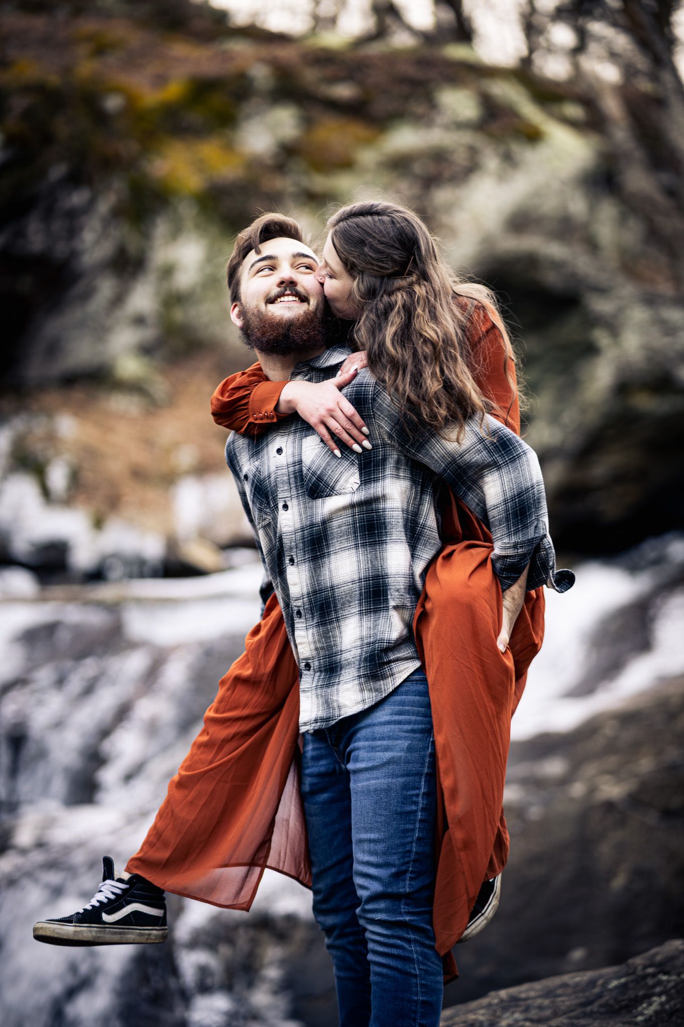 Man giving woman piggyback ride near waterfall as she kisses his cheek, both smiling joyfully.