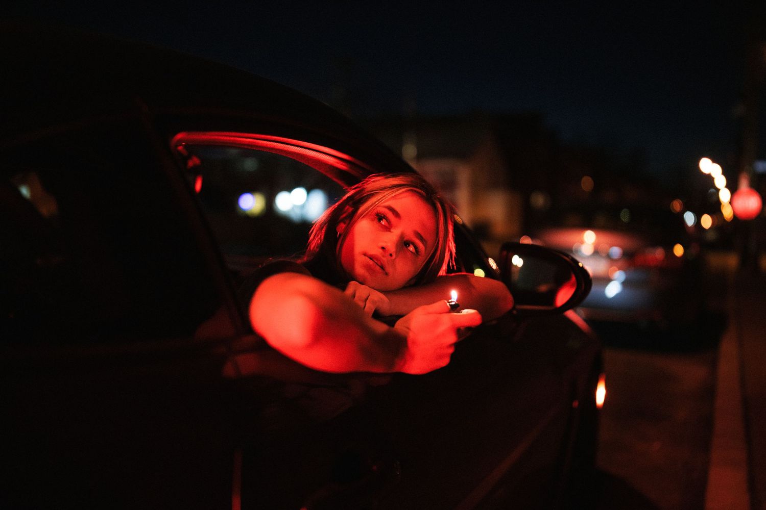 Woman leans out of car window lit by red neon glow on a dark city street at night.