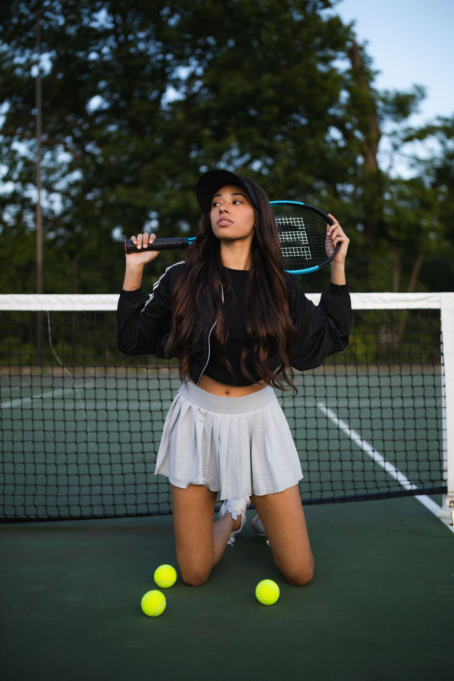 Young woman in black crop top and white pleated skirt kneeling on a tennis court holding a racket.