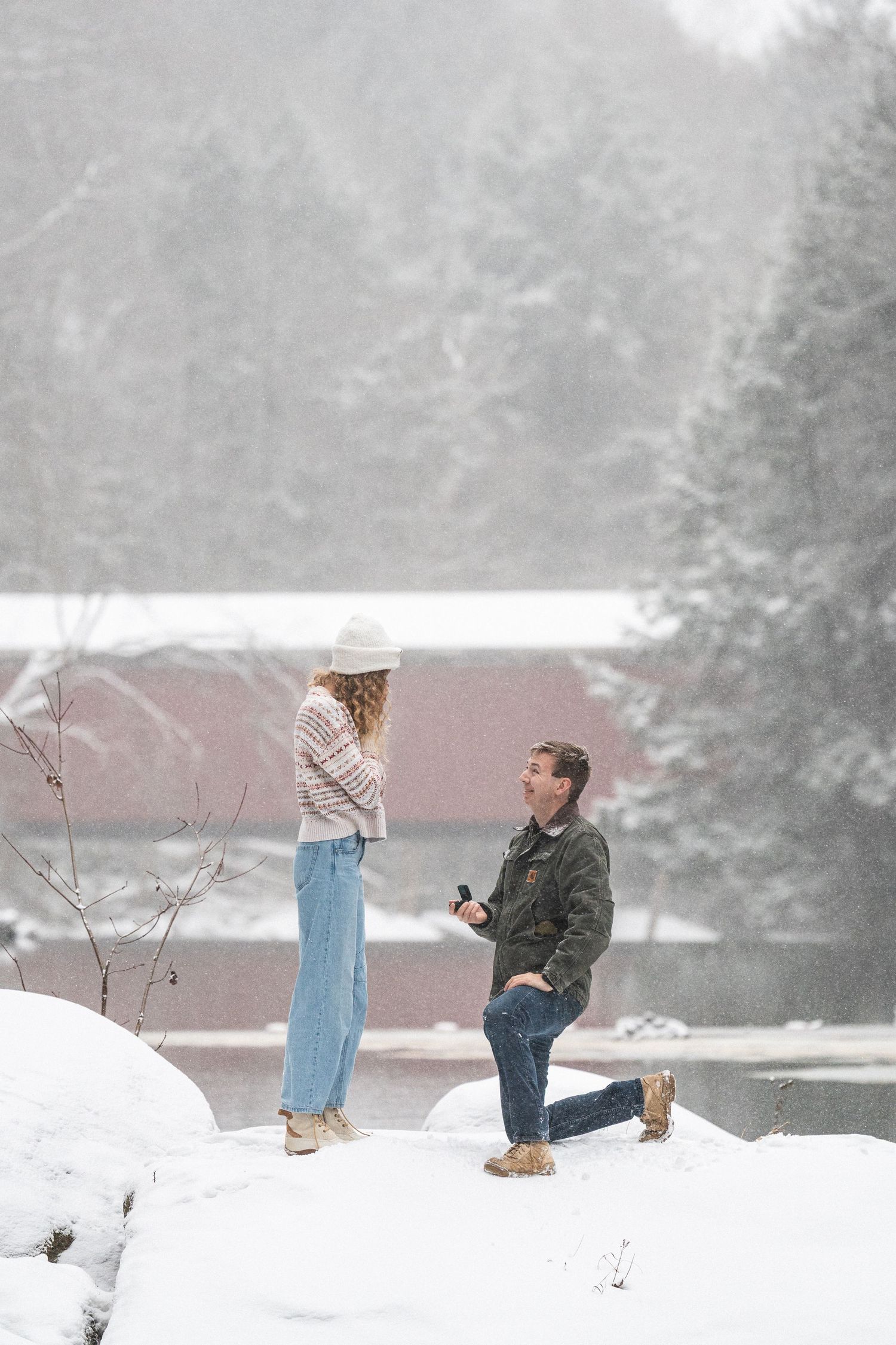 Man proposes to girlfriend on one knee in a snowy winter landscape near a red covered bridge.