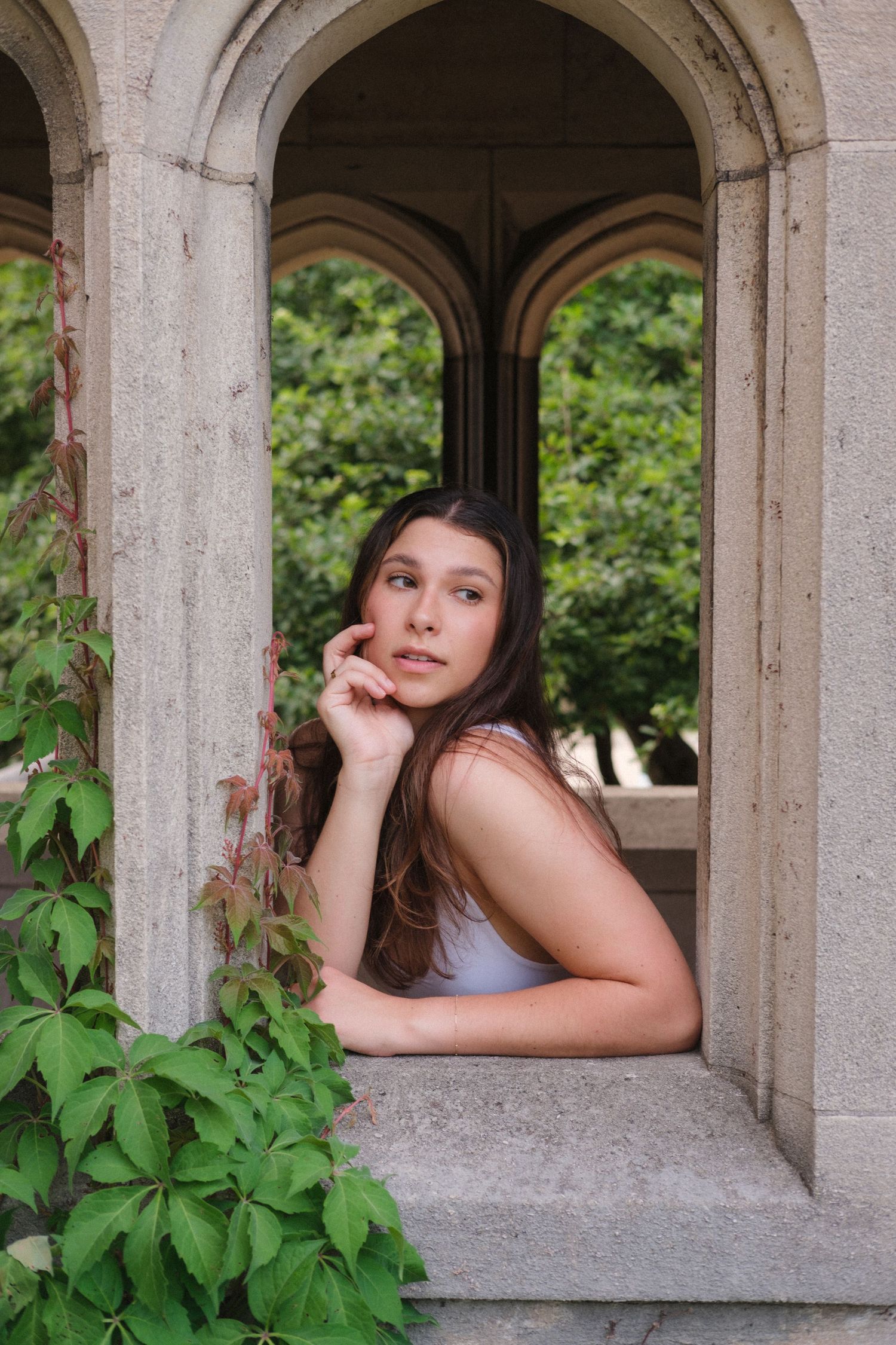 Young woman with long brown hair posing thoughtfully in a stone archway covered with green ivy.