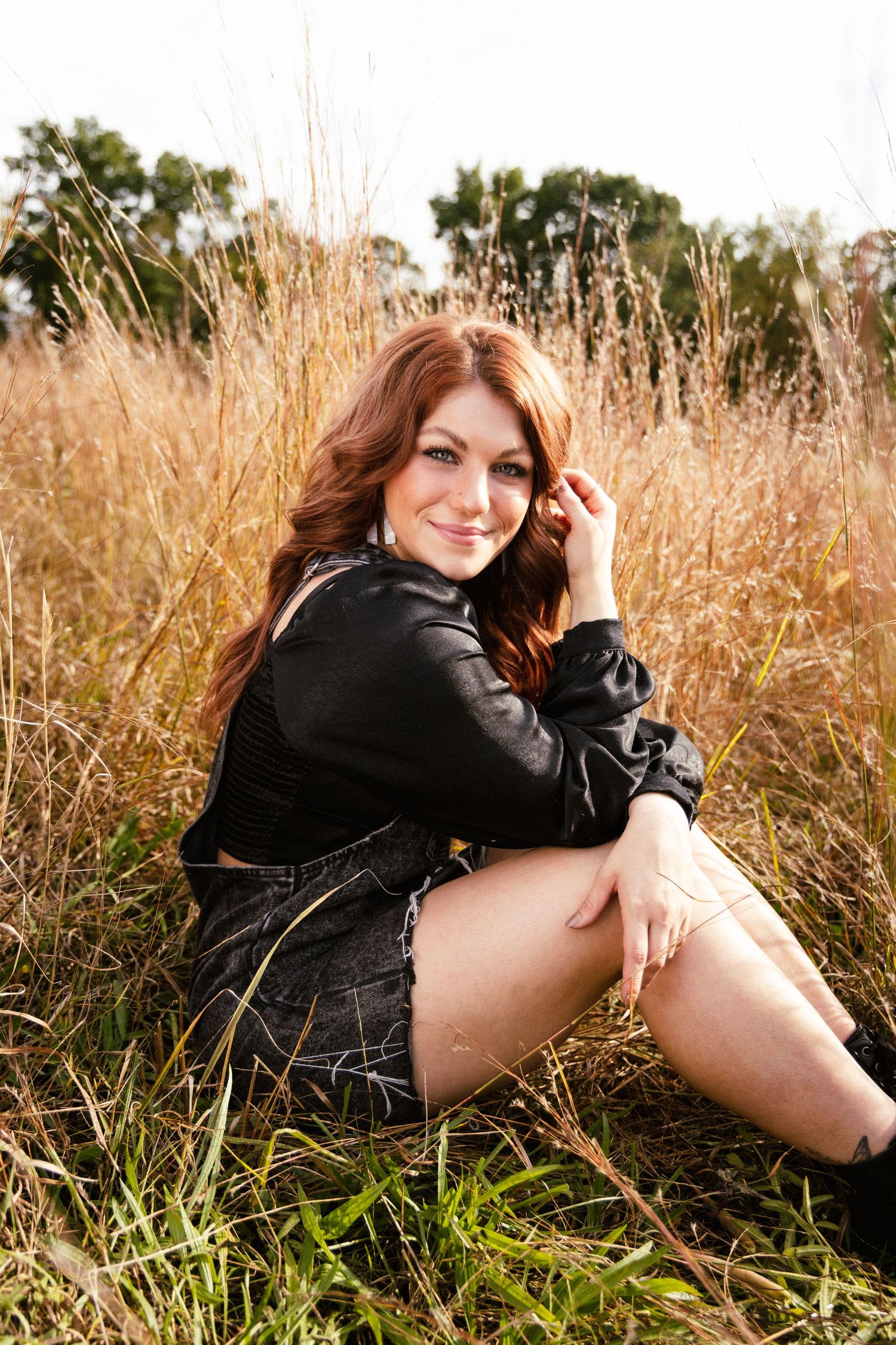 Young woman with red hair sits smiling in golden field grass wearing black outfit.