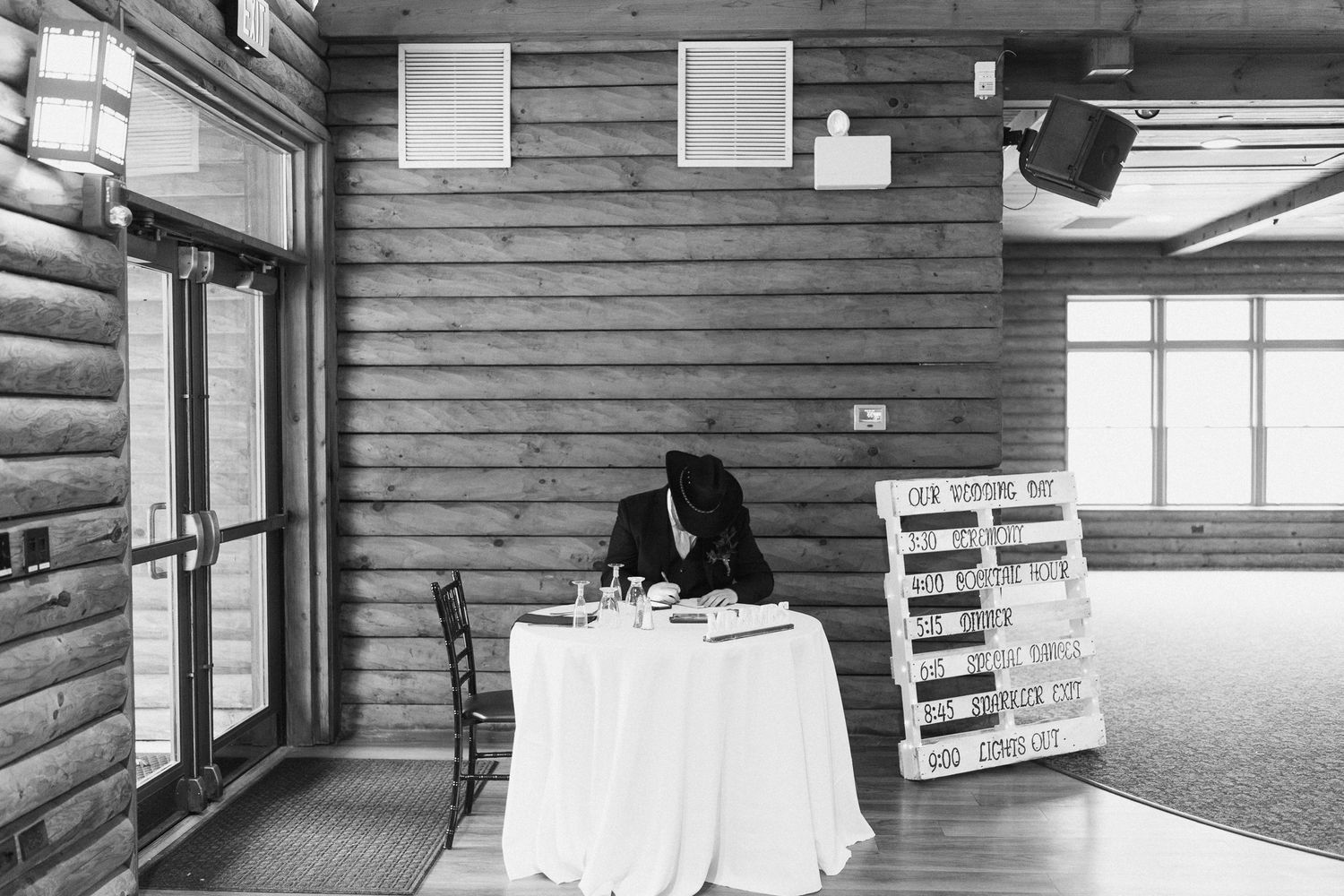 Black and white photo of a guest signing the wedding registry at a table in a log cabin reception hall.