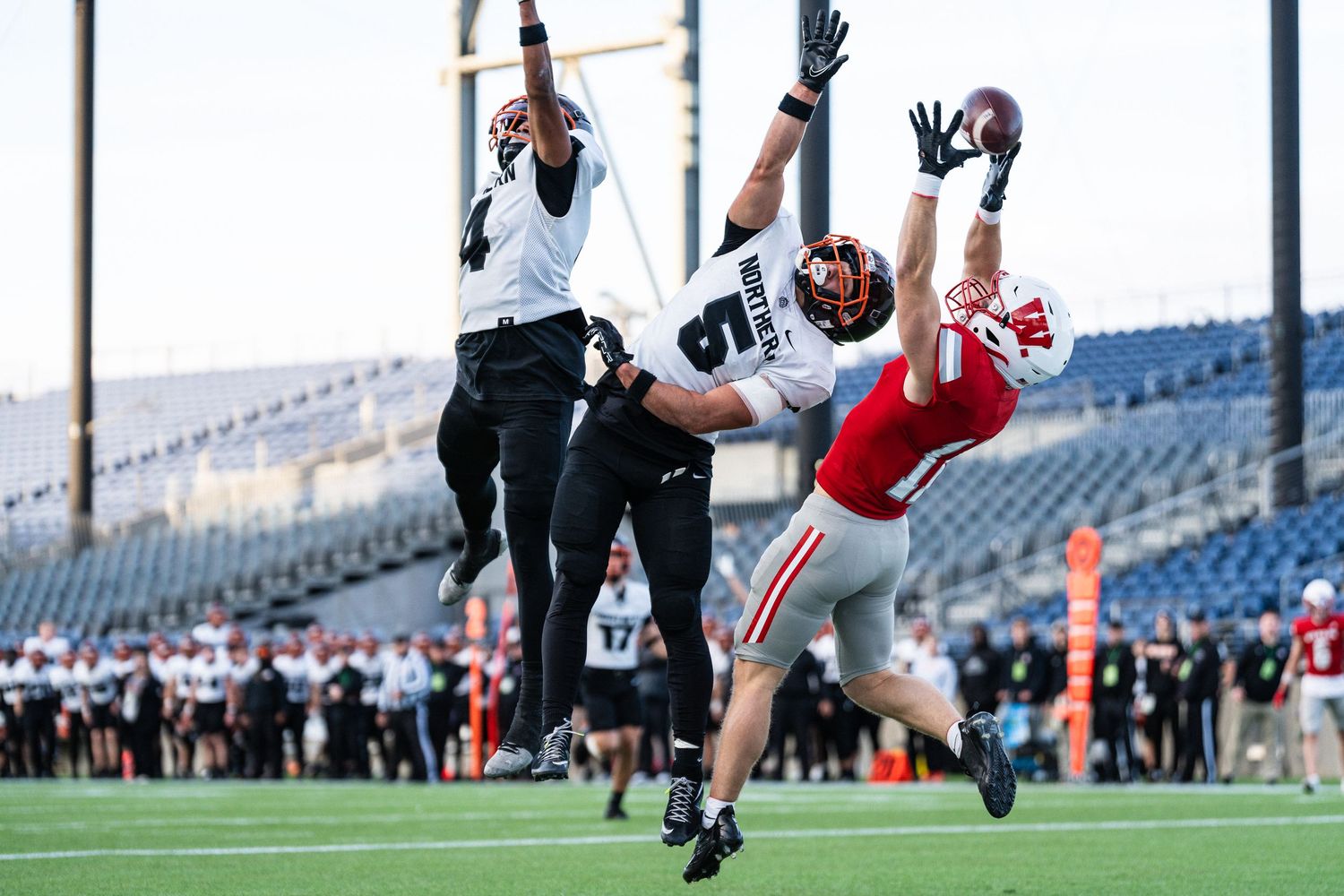 Two defenders in black uniforms leap to defend pass against red-uniformed receiver in stadium.