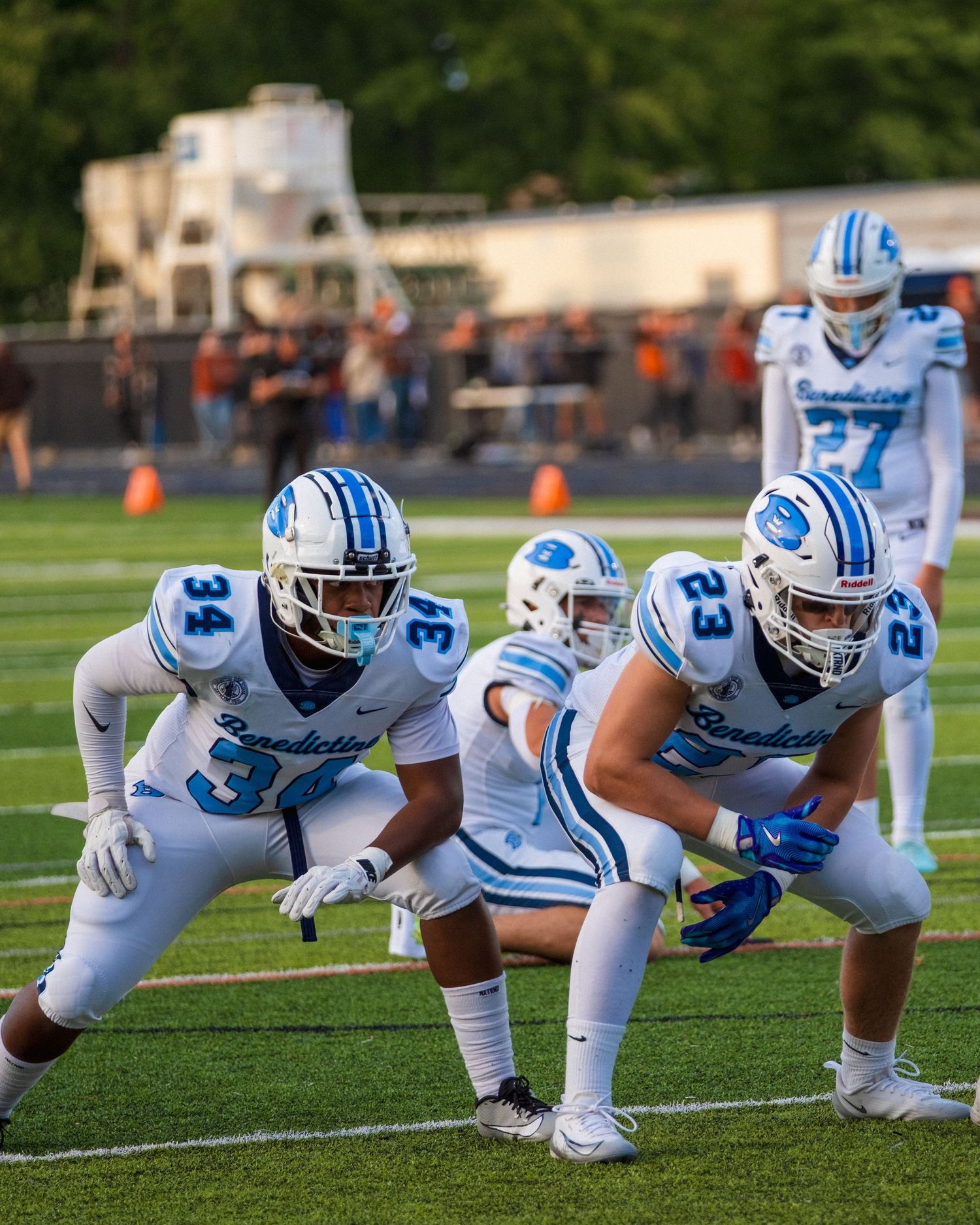 Benedictine football players in white and blue uniforms line up on the field during a game.