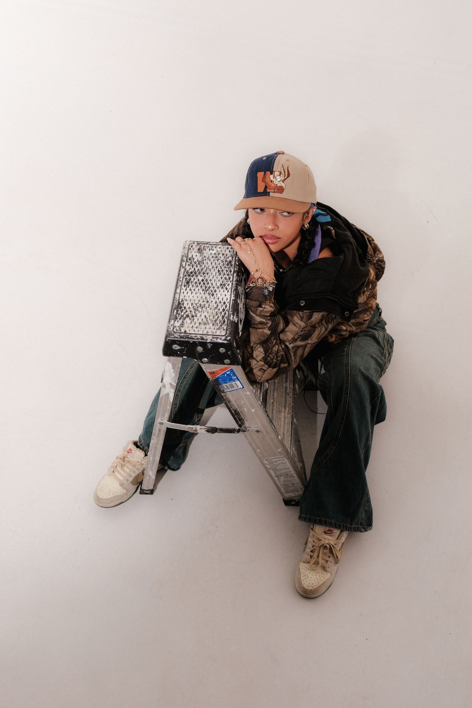 Young person in camo jacket and cap sits on ladder against white background.