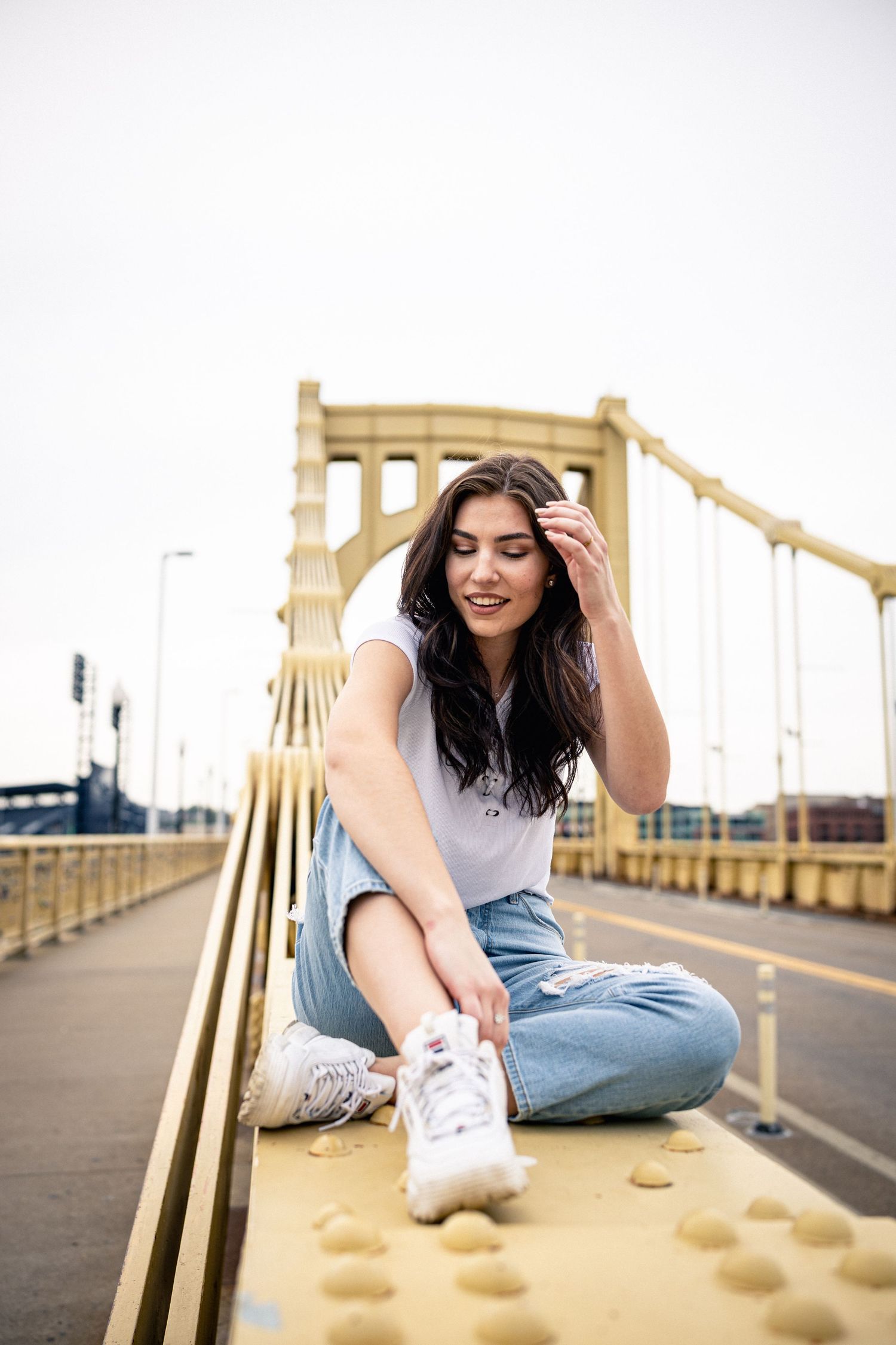 Brunette woman in white tee and ripped jeans sitting cross-legged on yellow bridge railing.