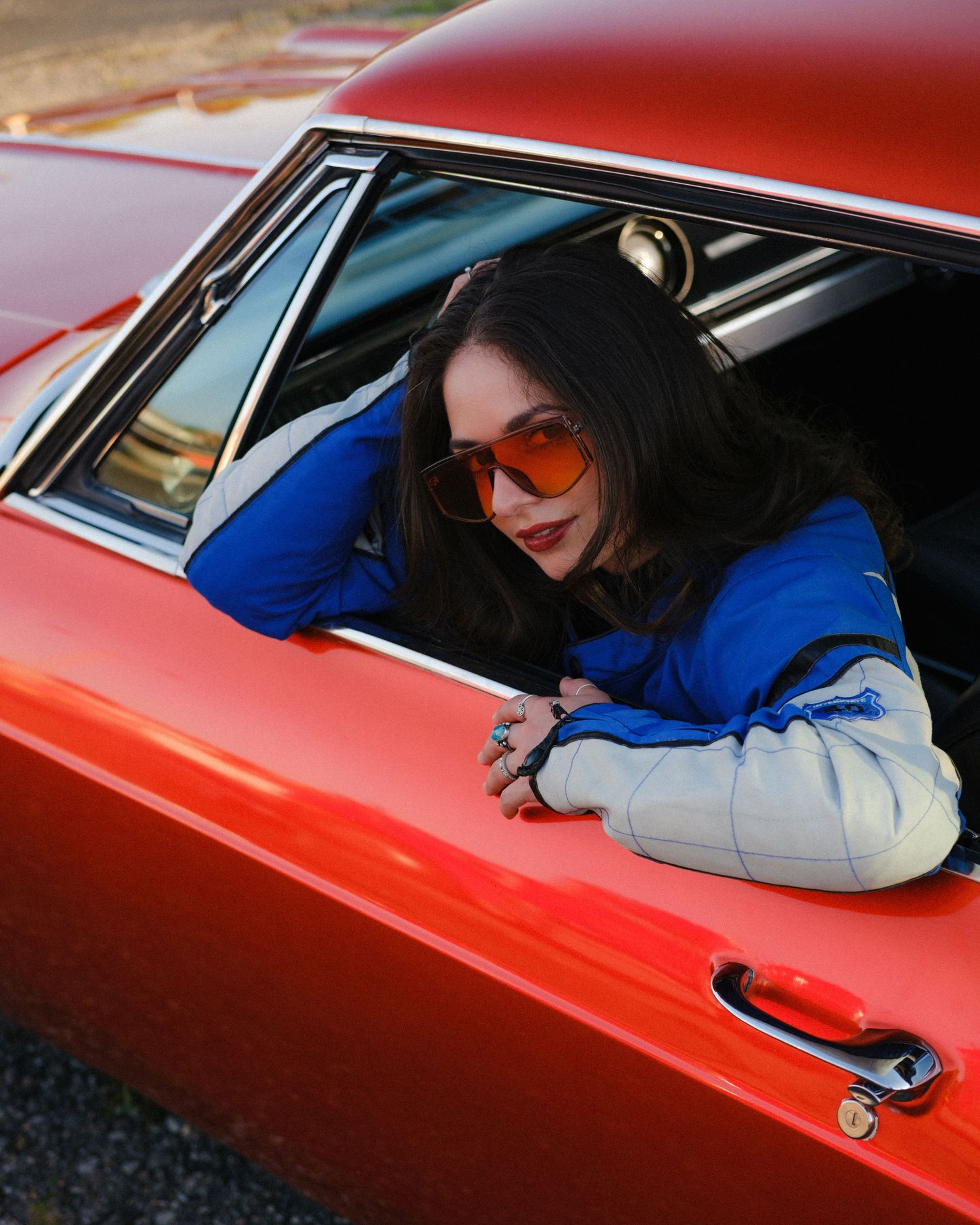 Young woman with dark hair and orange sunglasses leaning out of a classic red car window.