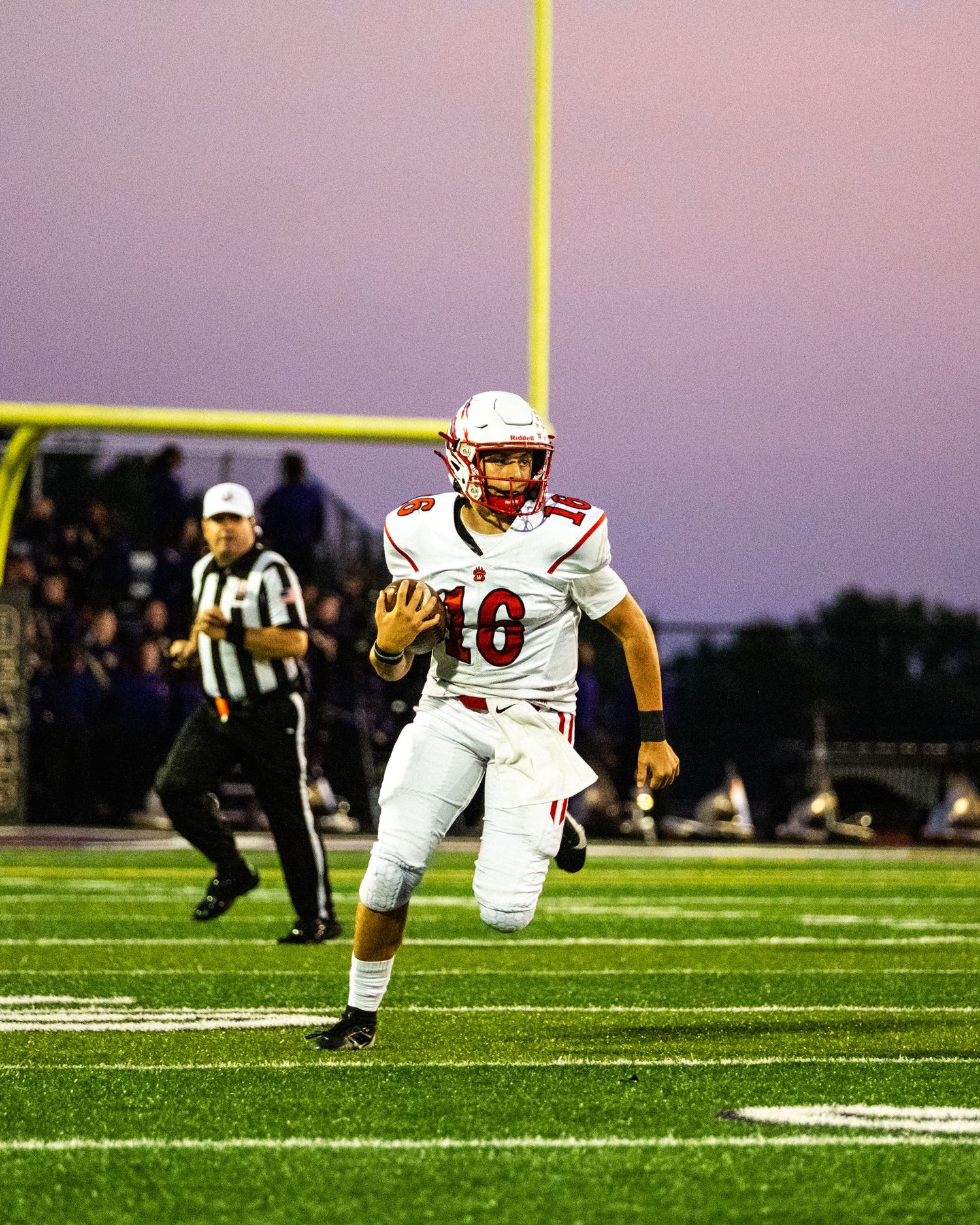 High school football player wearing number 10 red jersey runs with ball on green field at dusk.