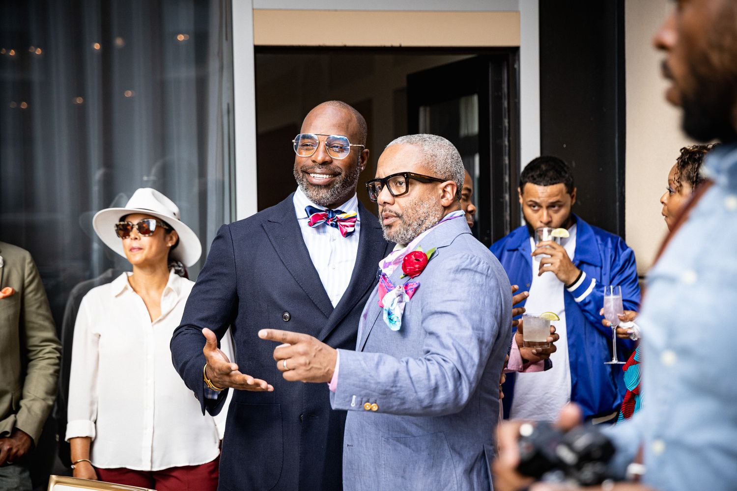 Two well-dressed men in colorful bow ties converse at an outdoor social gathering with guests in background.