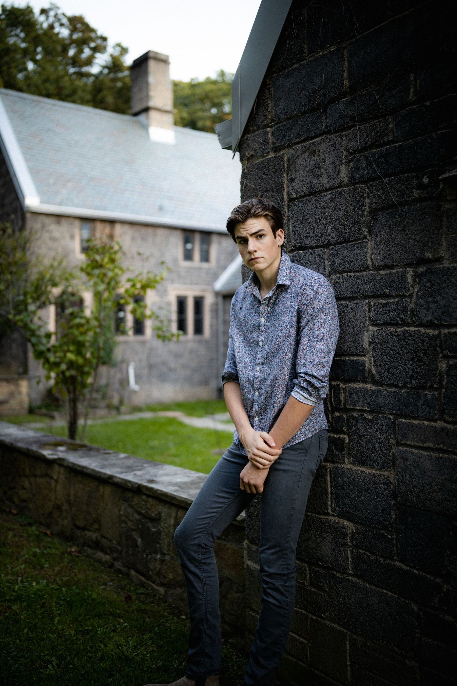 Young man in floral shirt leaning against dark brick wall with historic stone building in background.