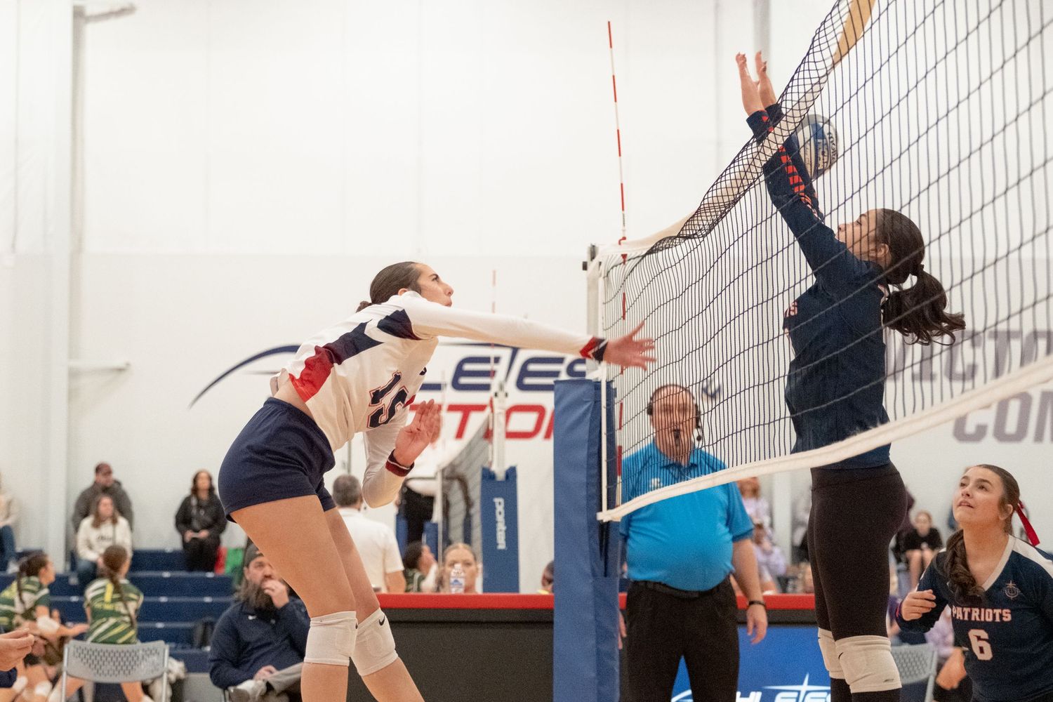 Female volleyball players compete at the net during an indoor match with spectators watching.