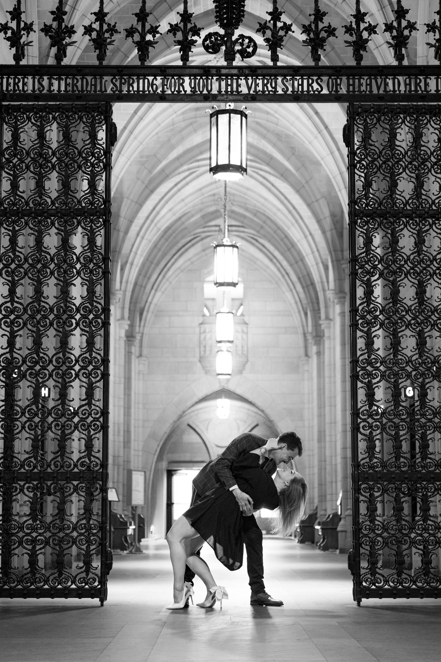 Black and white romantic dip kiss framed by ornate iron gates inside a grand Gothic archway.