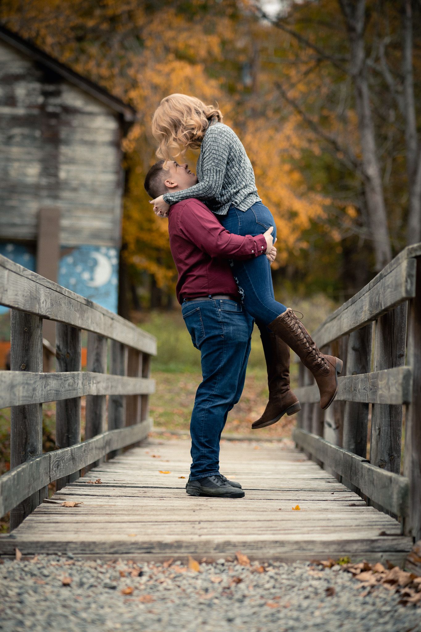 Man lifting his girlfriend joyfully on a rustic wooden bridge during an autumn engagement session.