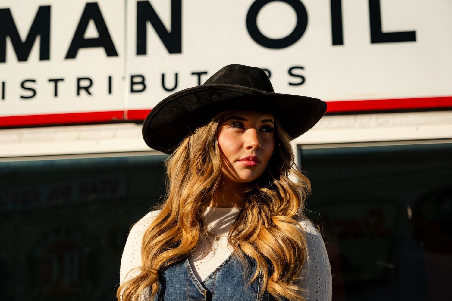 Stylish woman in black wide-brim hat and denim vest standing before a vintage Man Oil Distributors sign.