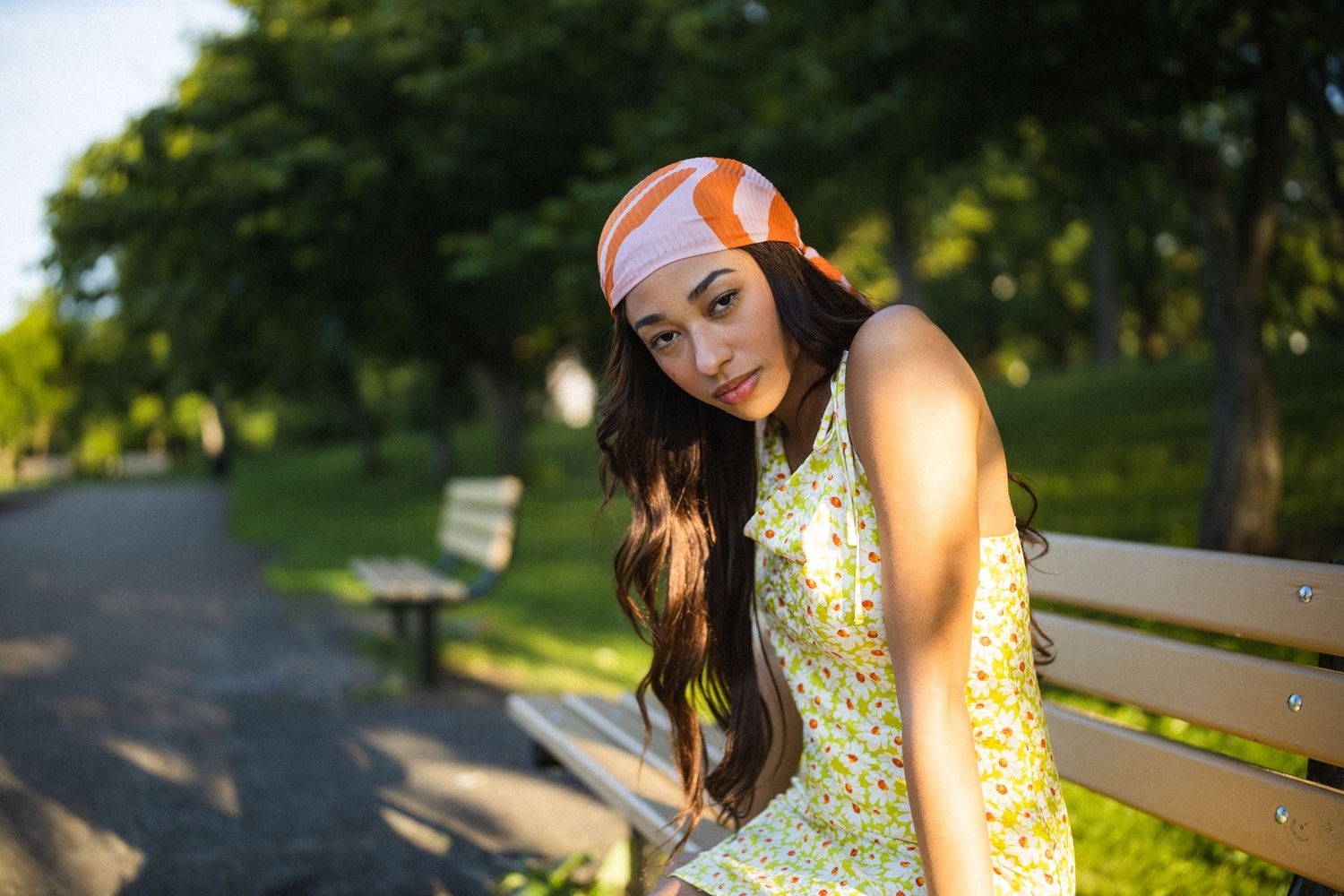 Young woman in floral dress and orange headband sits on park bench surrounded by green trees.
