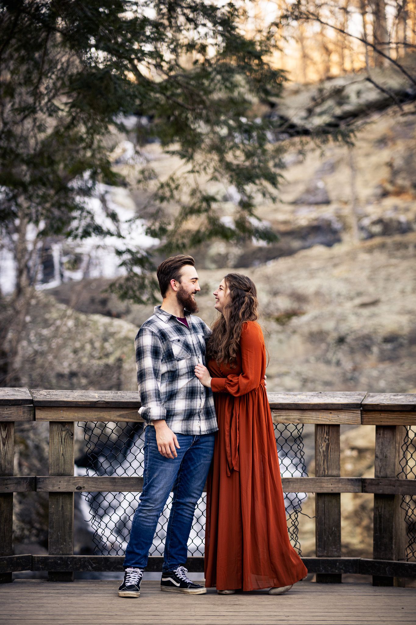 Romantic couple in casual attire gazing at each other on wooden bridge surrounded by nature.
