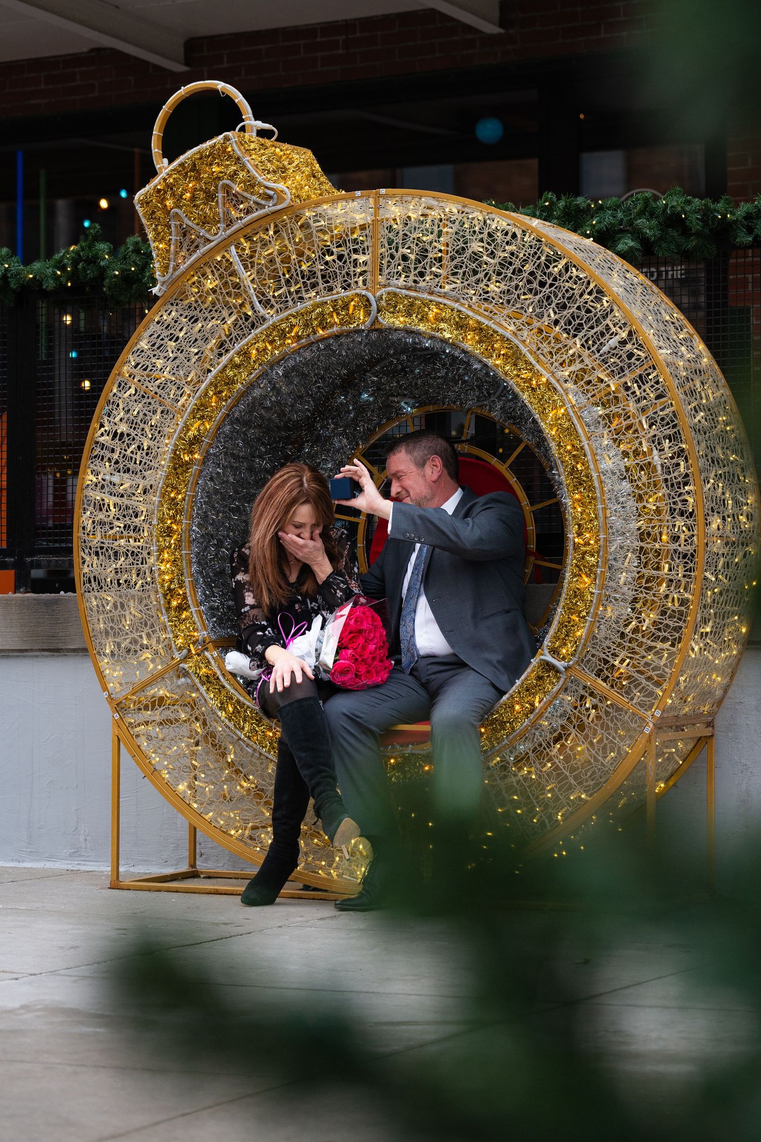 Couple sits inside a large illuminated gold Christmas ornament decoration taking photos together.