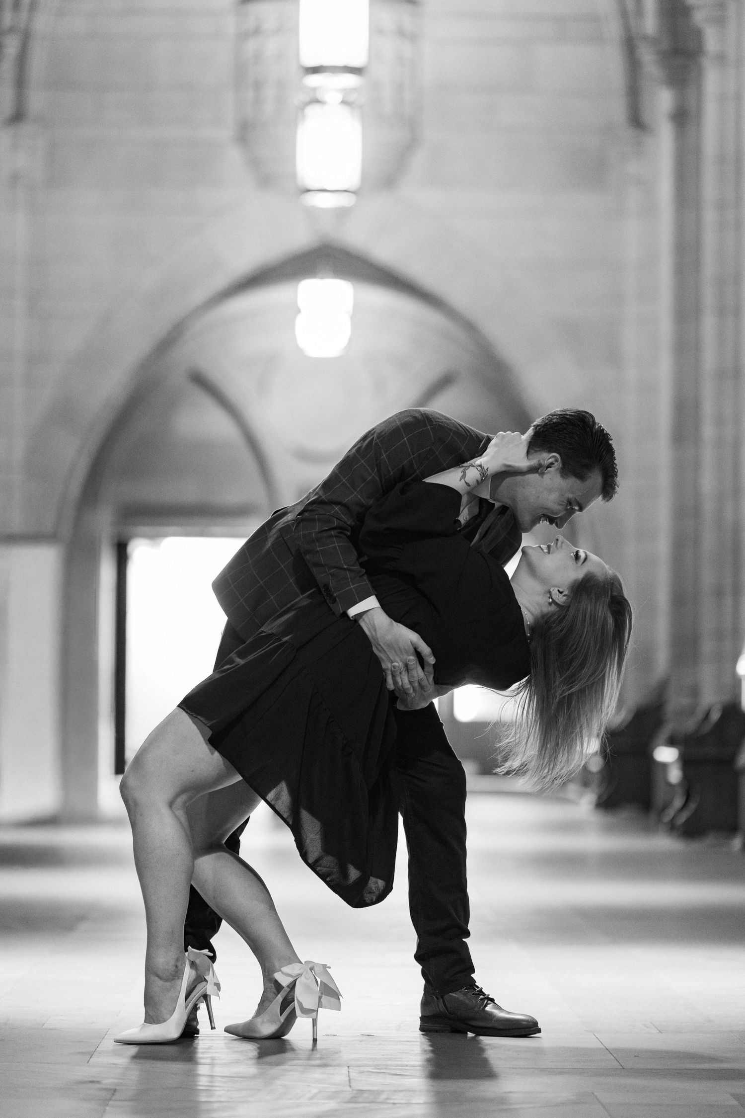 Black and white photo of a couple sharing a romantic dip kiss inside an elegant arched stone corridor.