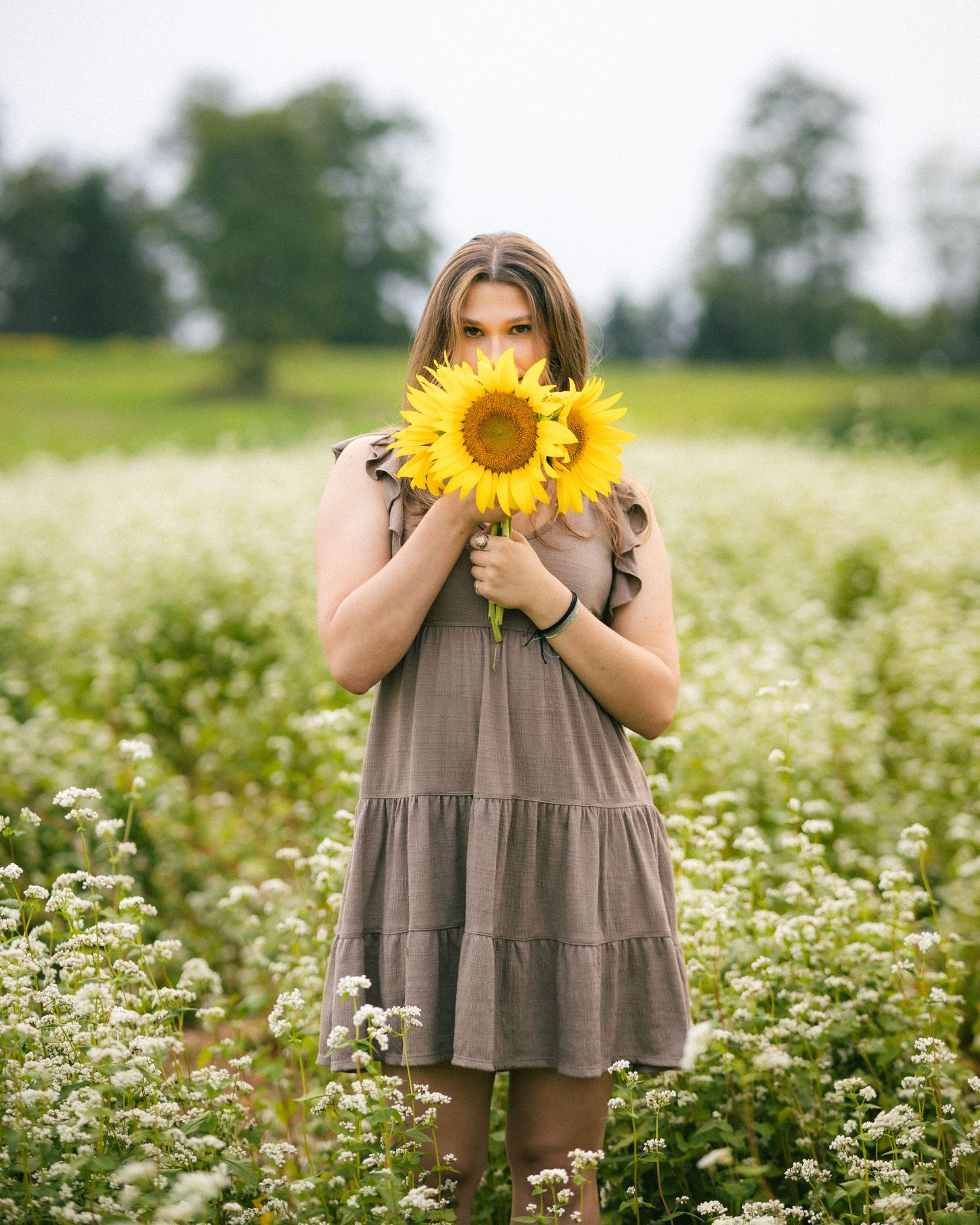 Young woman in gray dress holding bright sunflowers covering her face in a white wildflower field.