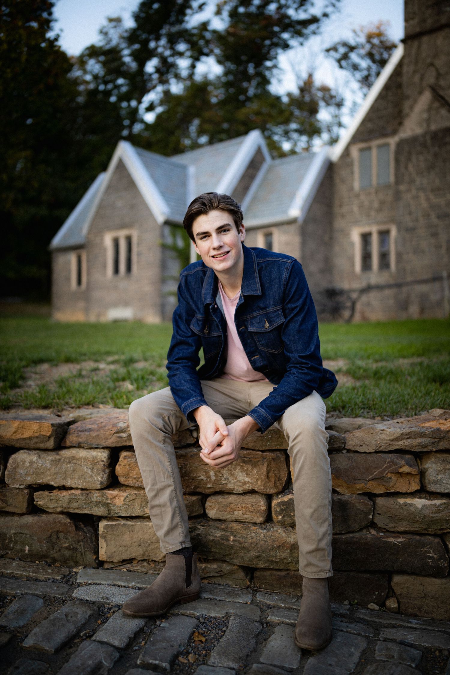 Smiling young man in denim jacket sitting on stone wall with historic stone chapel in background.