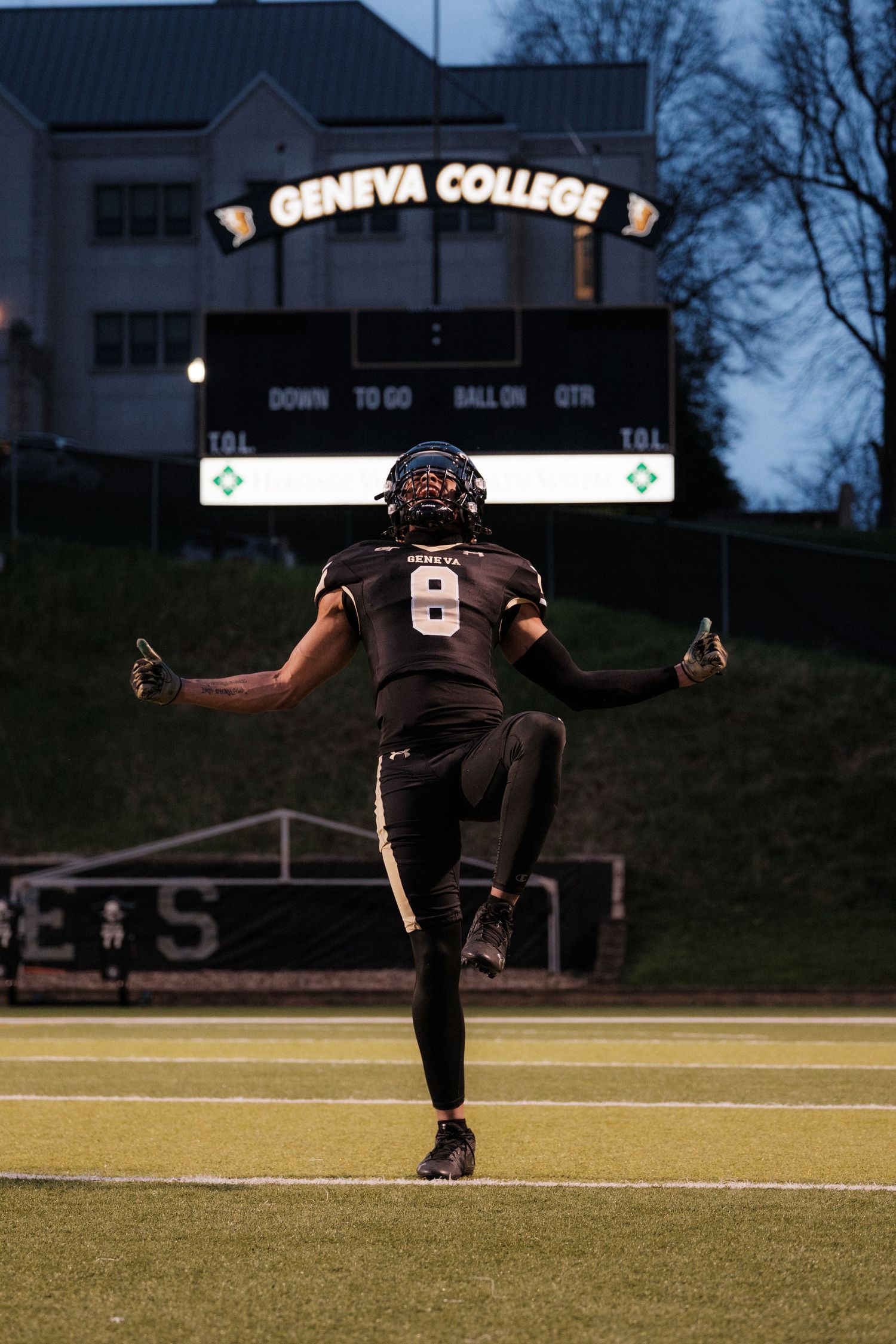 Geneva College football player number 8 celebrates on field with scoreboard lit at night.