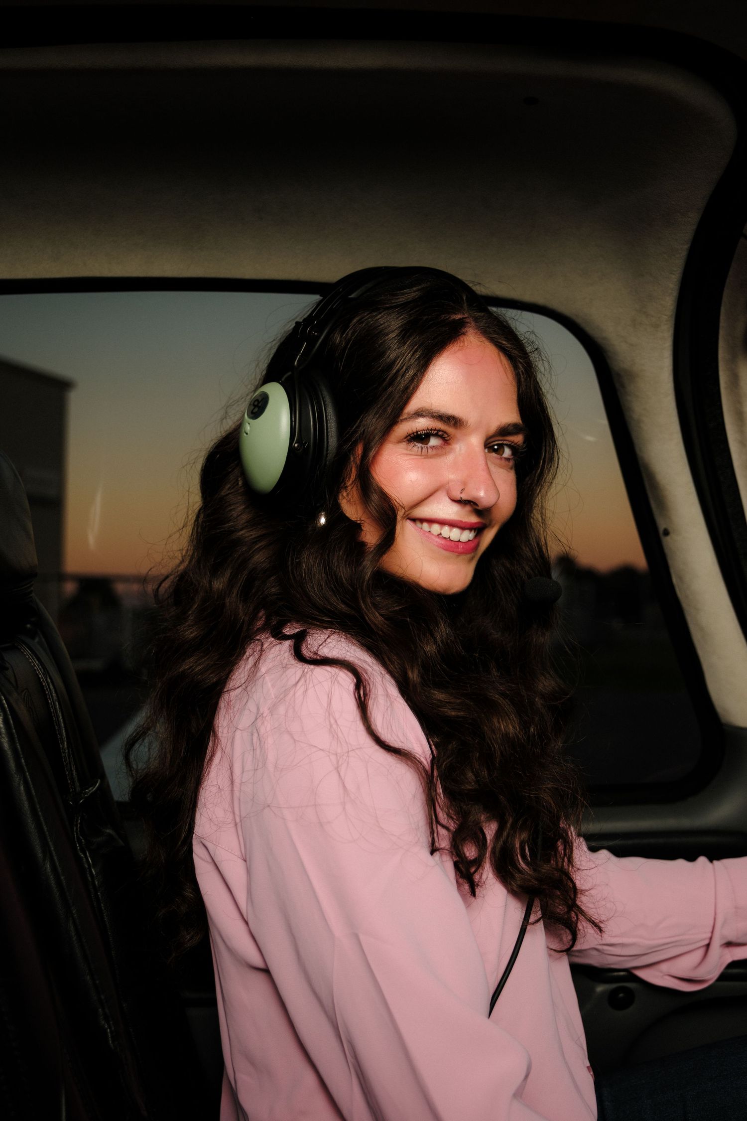 Smiling woman with curly hair wearing green headphones inside aircraft cockpit at sunset.