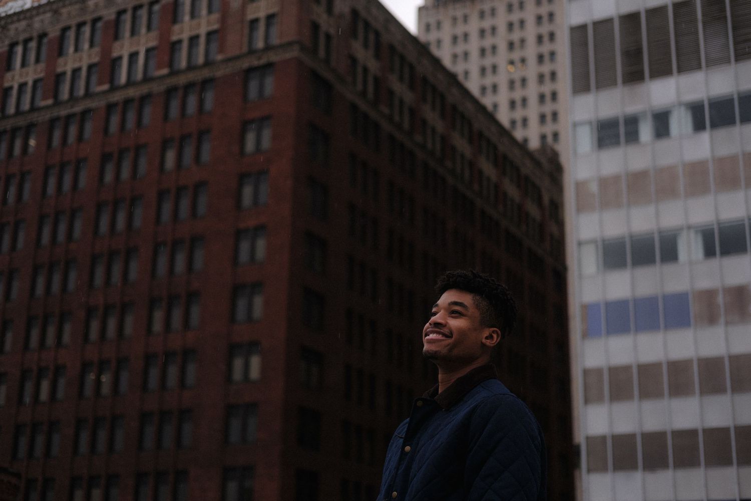 Young Black man smiling confidently outdoors with urban brick buildings in the background on an overcast day.