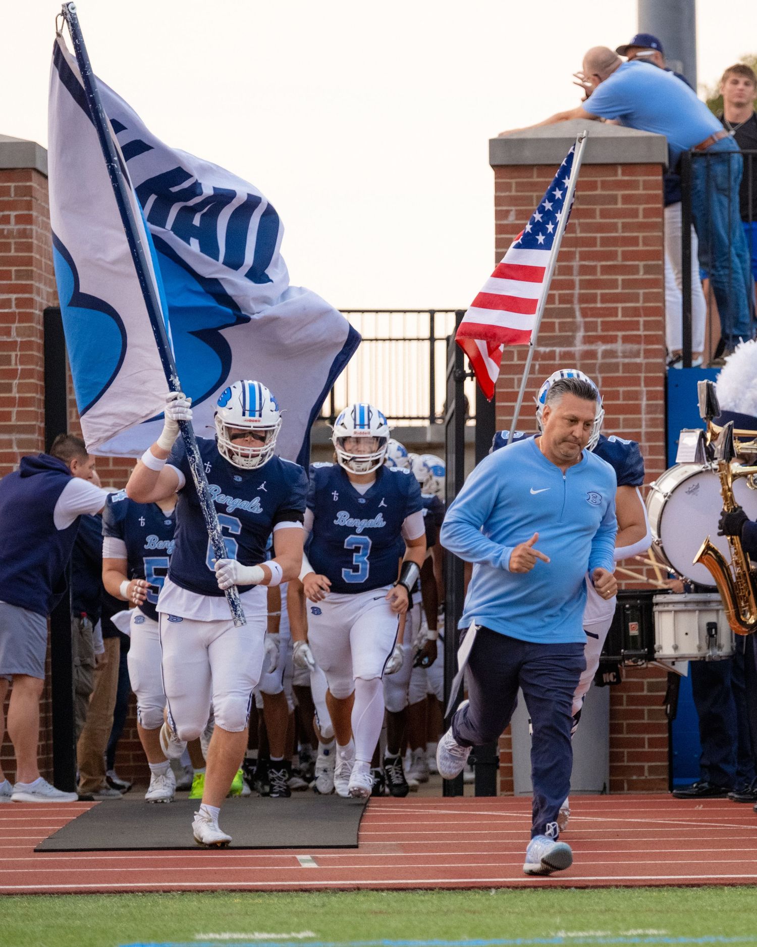 Benedictine football team runs onto the field with flags and coach leading the way.