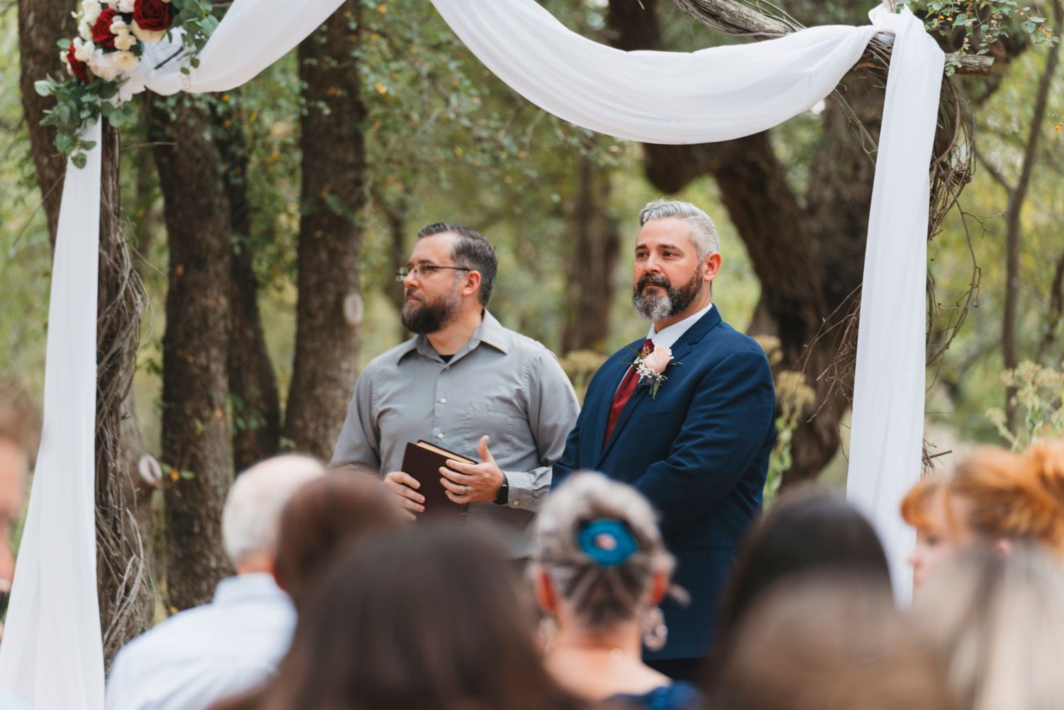 Groom in navy suit stands at outdoor woodland wedding altar with officiant under white draped arch.
