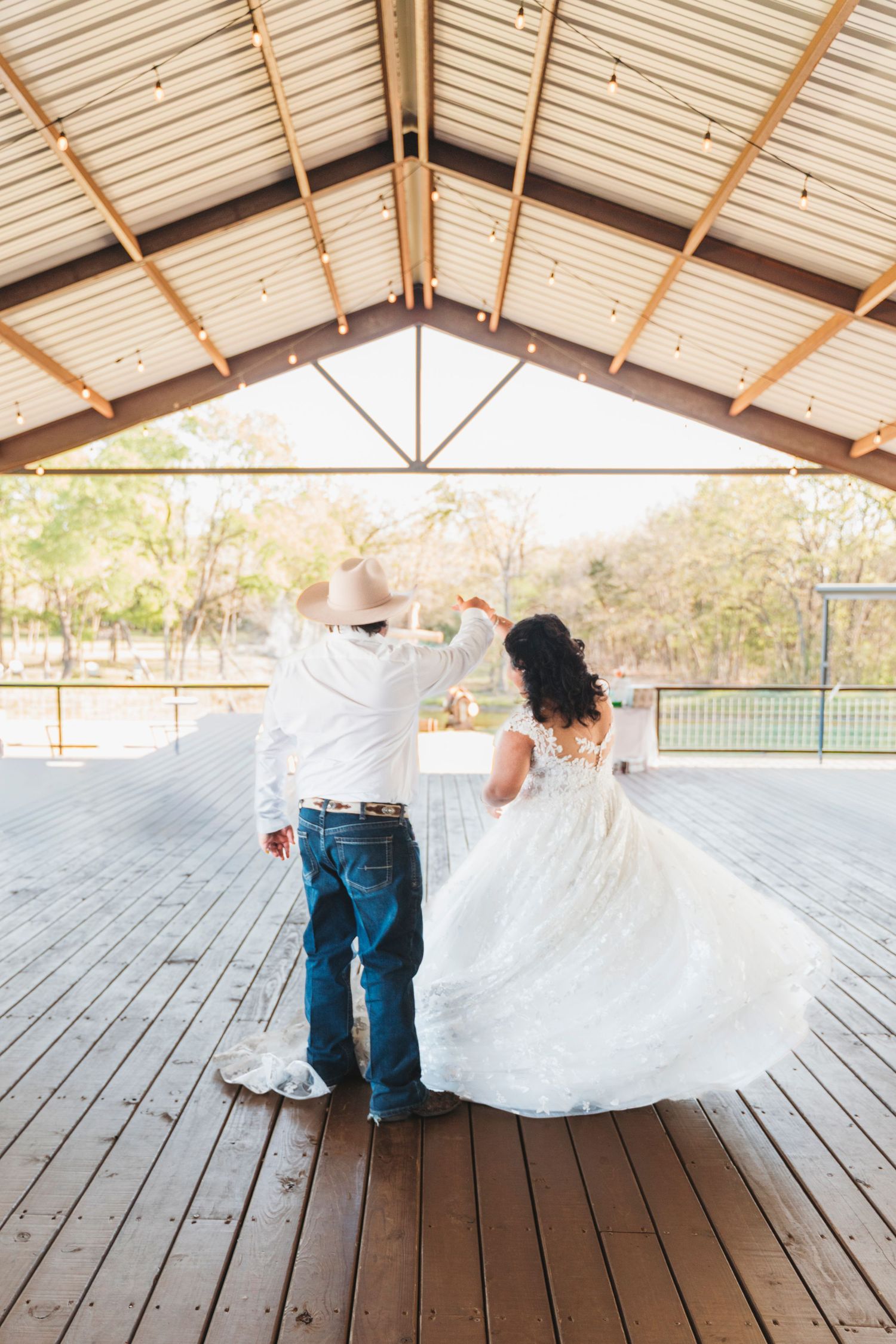 Bride and groom dancing under rustic pavilion, bride in flowing white gown, groom in cowboy hat and jeans.