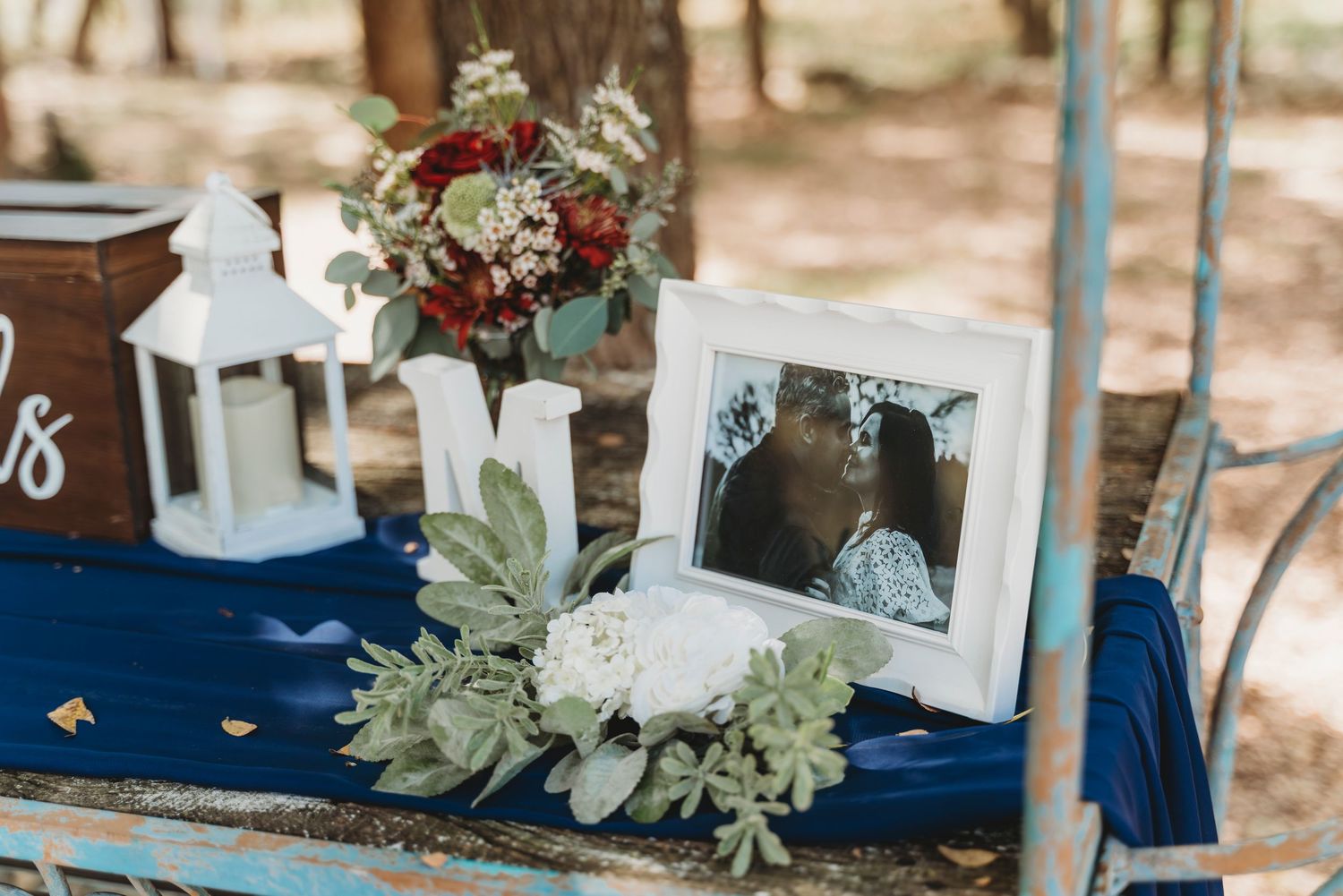 Rustic wedding table with framed couple photo, white lantern, floral bouquet, and navy blue linen decor outdoors.