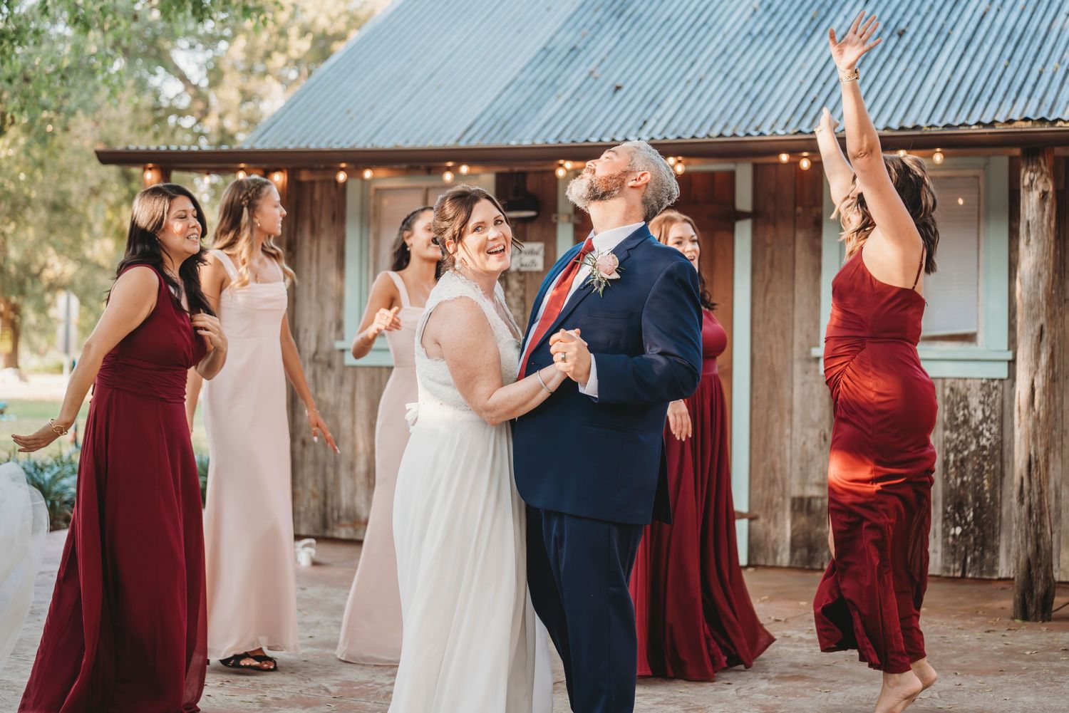 Bride and groom share first dance outdoors while bridesmaids in red and blush gowns celebrate at rustic barn wedding.
