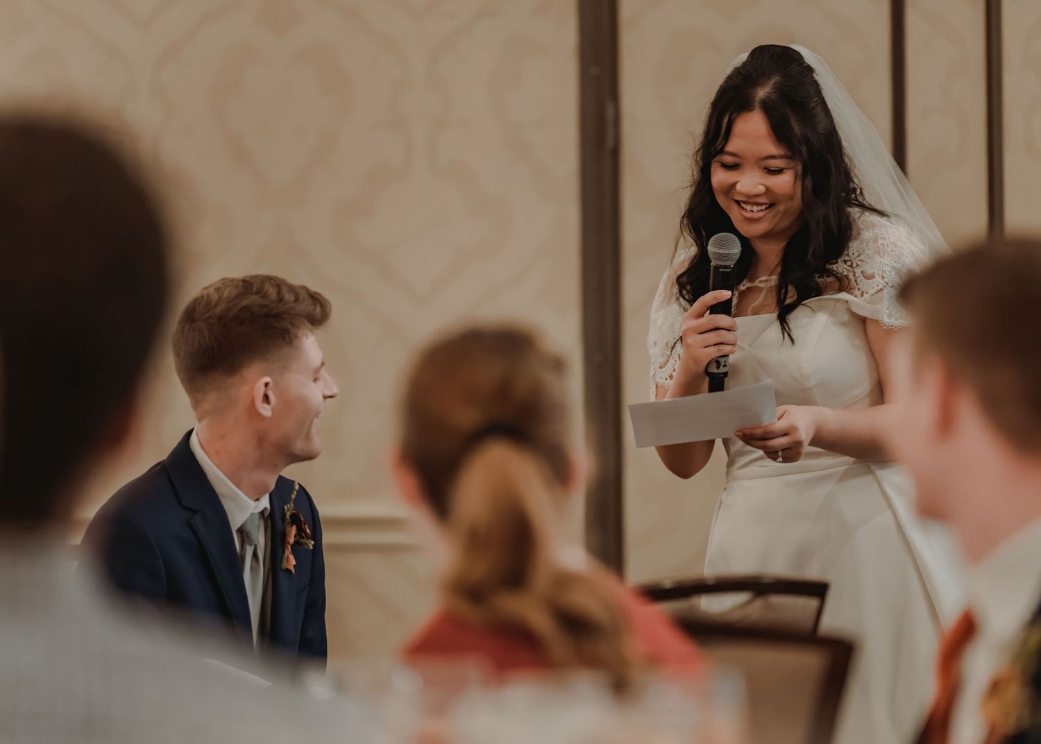 Bride gives a heartfelt speech at wedding reception, smiling while holding microphone and notes.