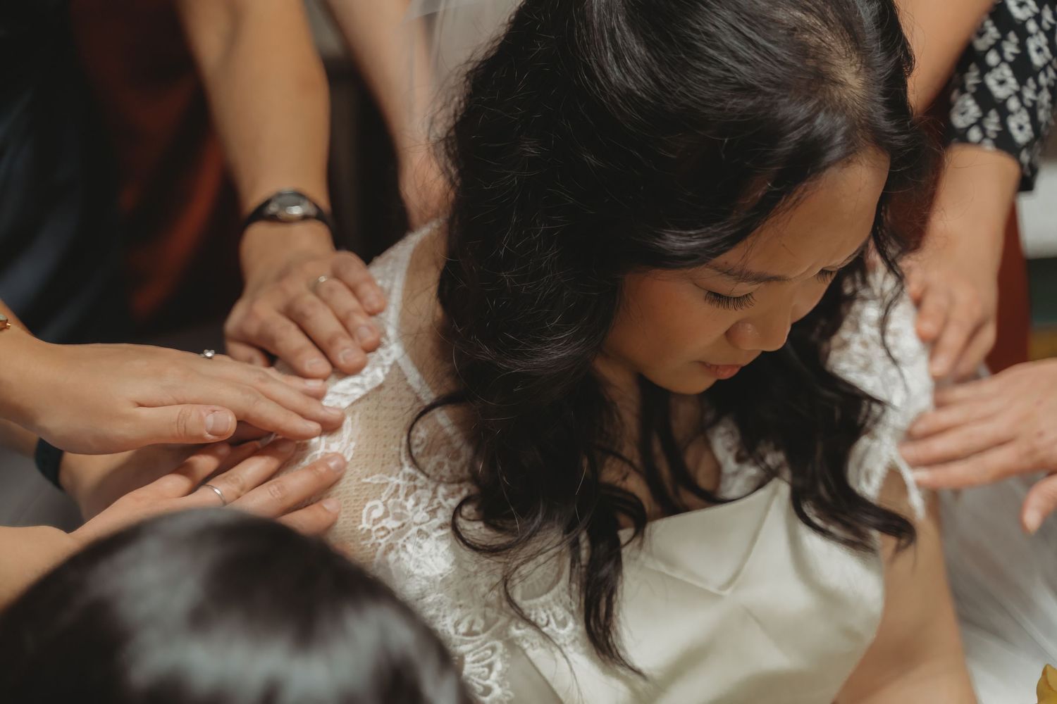 Bride in lace wedding dress surrounded by hands helping her prepare on her wedding day.