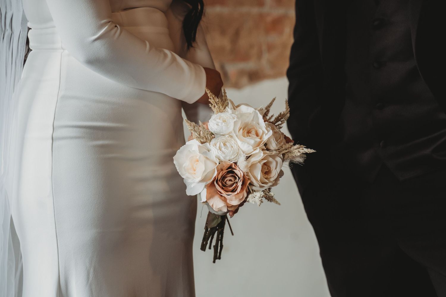 Bride in white gown holding elegant bouquet of white and rose gold flowers beside groom in black suit.