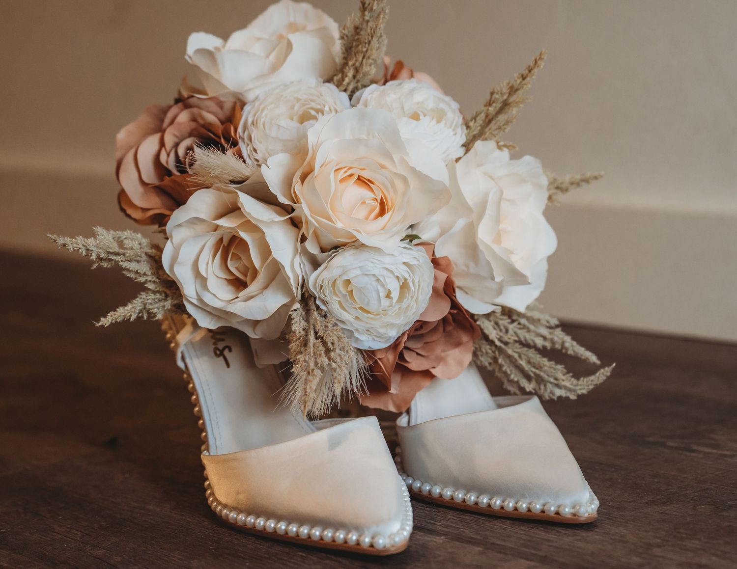 Elegant bridal heels adorned with pearls beside a romantic bouquet of white and blush roses with dried pampas grass.