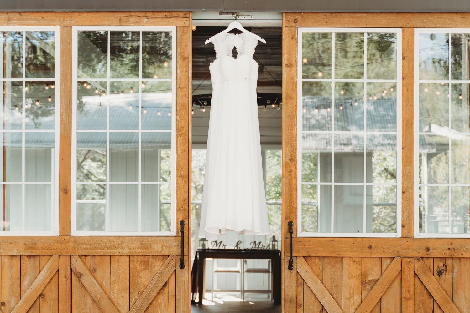 White wedding dress hanging on barn doors with large windows and lush greenery backdrop.