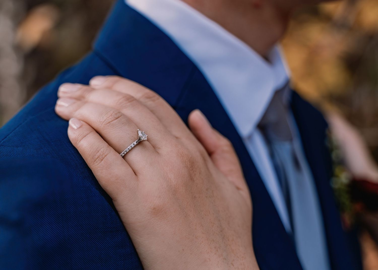 Close-up of a woman's hand with a diamond engagement ring resting on a man's navy blue suit jacket.