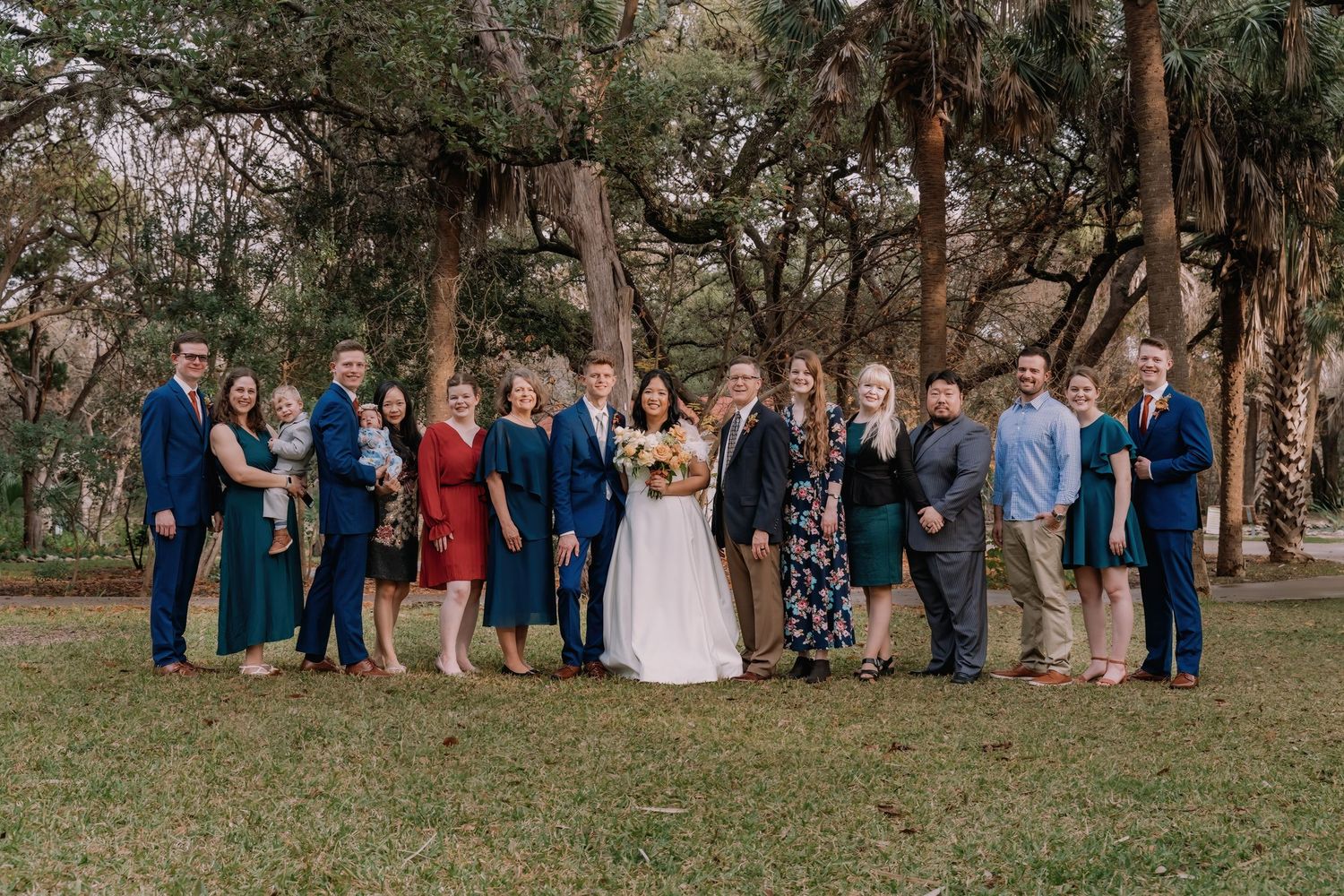 Large wedding party posing outdoors under sprawling oak trees, bride in white gown surrounded by family in formal attire.