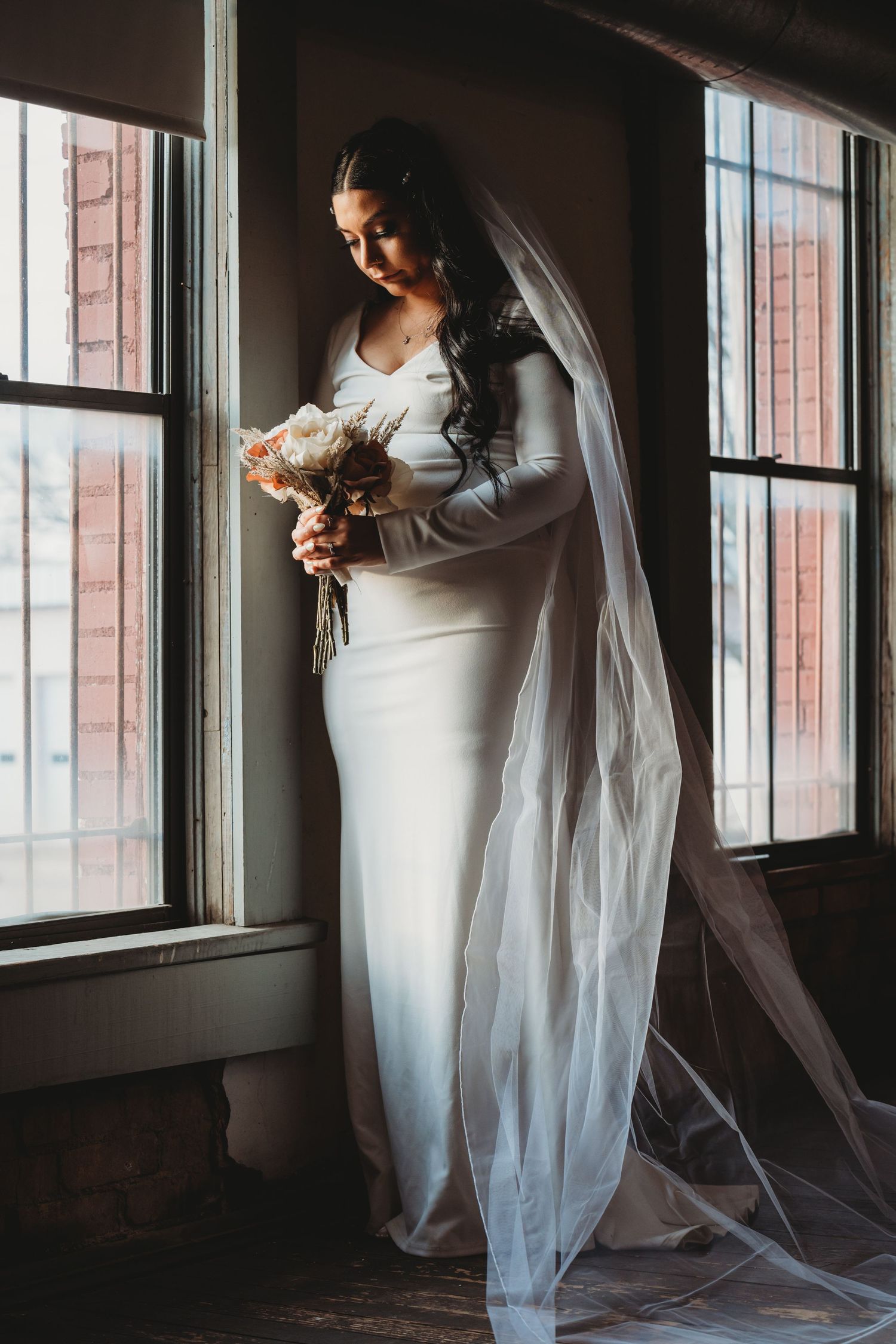 Bride in white long-sleeve gown with flowing veil holds bouquet by window in moody indoor setting.