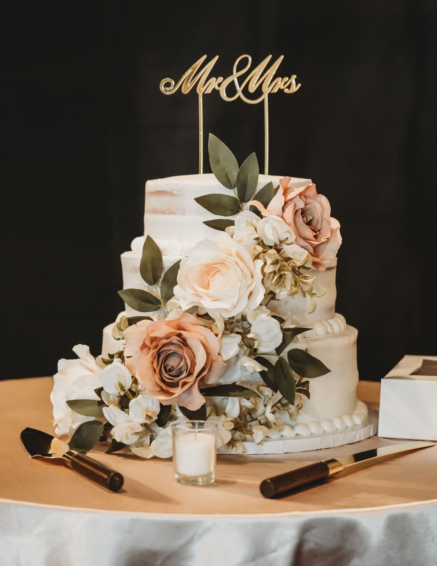 Two-tier white wedding cake with blush roses, greenery, and a gold Mr & Mrs topper on a elegant table setting.