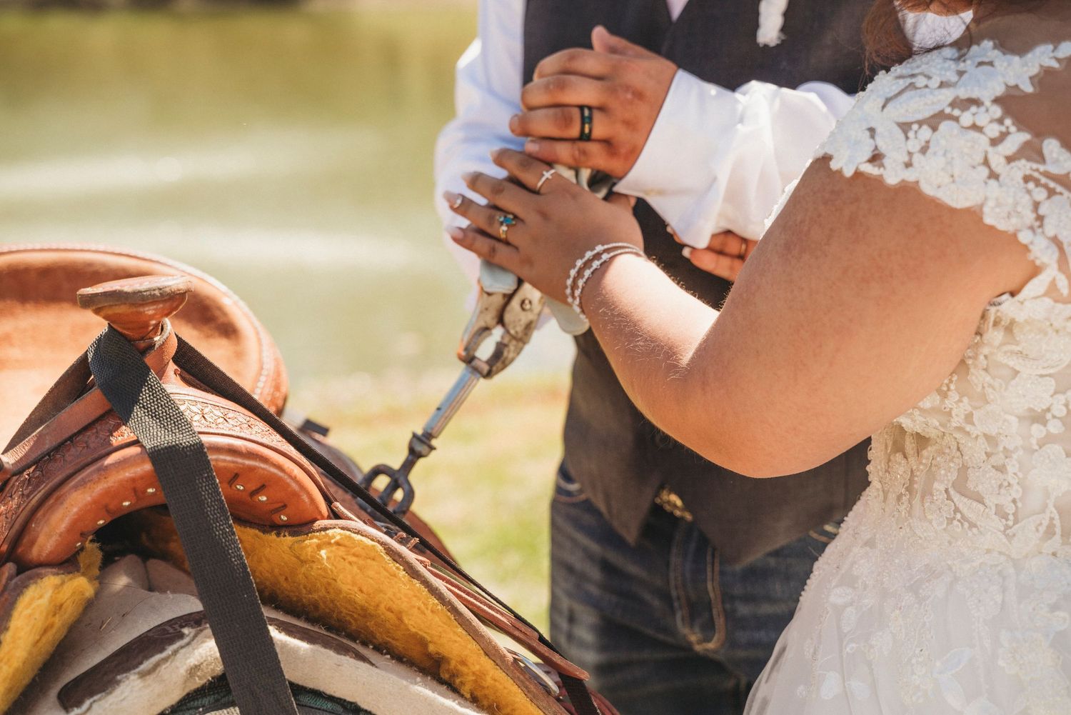 Bride and groom holding hands near a western saddle outdoors, bride wearing lace dress and pearl bracelet.