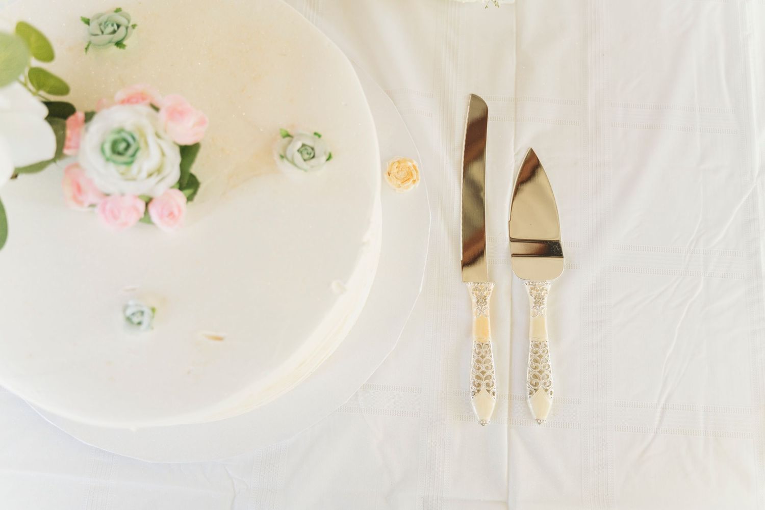 White wedding cake with pink roses and succulents beside gold cake knife and server on white linen.